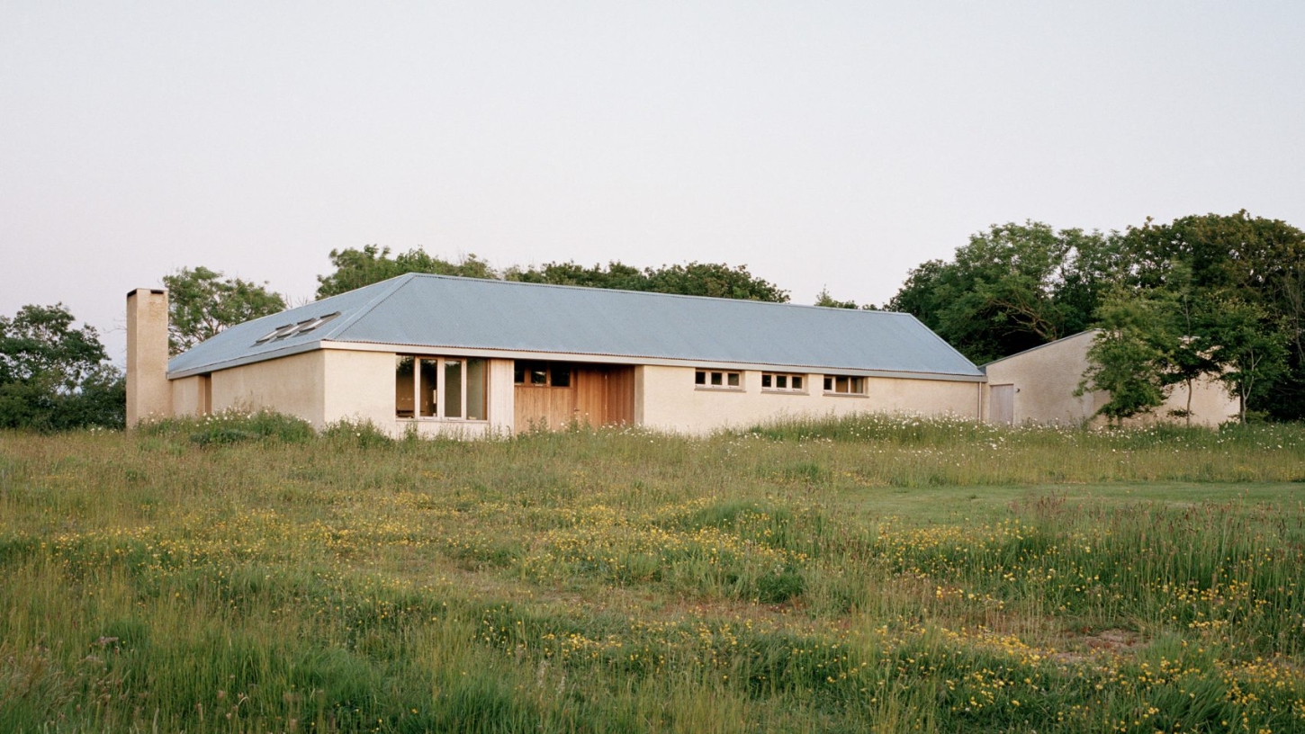 Farmworker's House by Hugh Strange Architects — A rendered farmhouse with cream-colored walls and dark standing-seam metal roof sits low within a wildflower meadow dotted with yellow blooms and tall grasses.