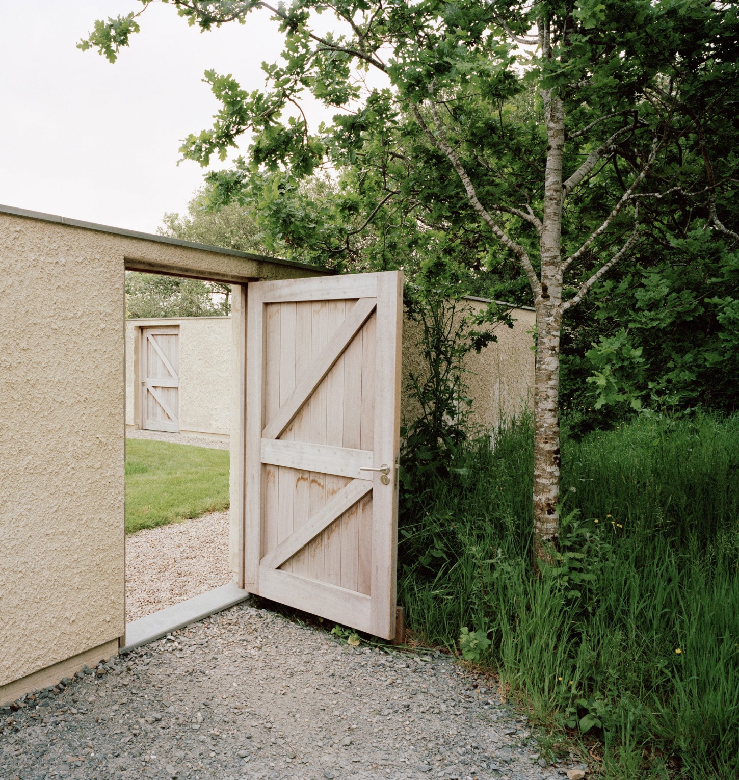 Farmworker's House by Hugh Strange Architects — A rendered timber-framed door stands open within a cream-colored rendered gateway structure, revealing a gravel courtyard and distant meadow beyond.