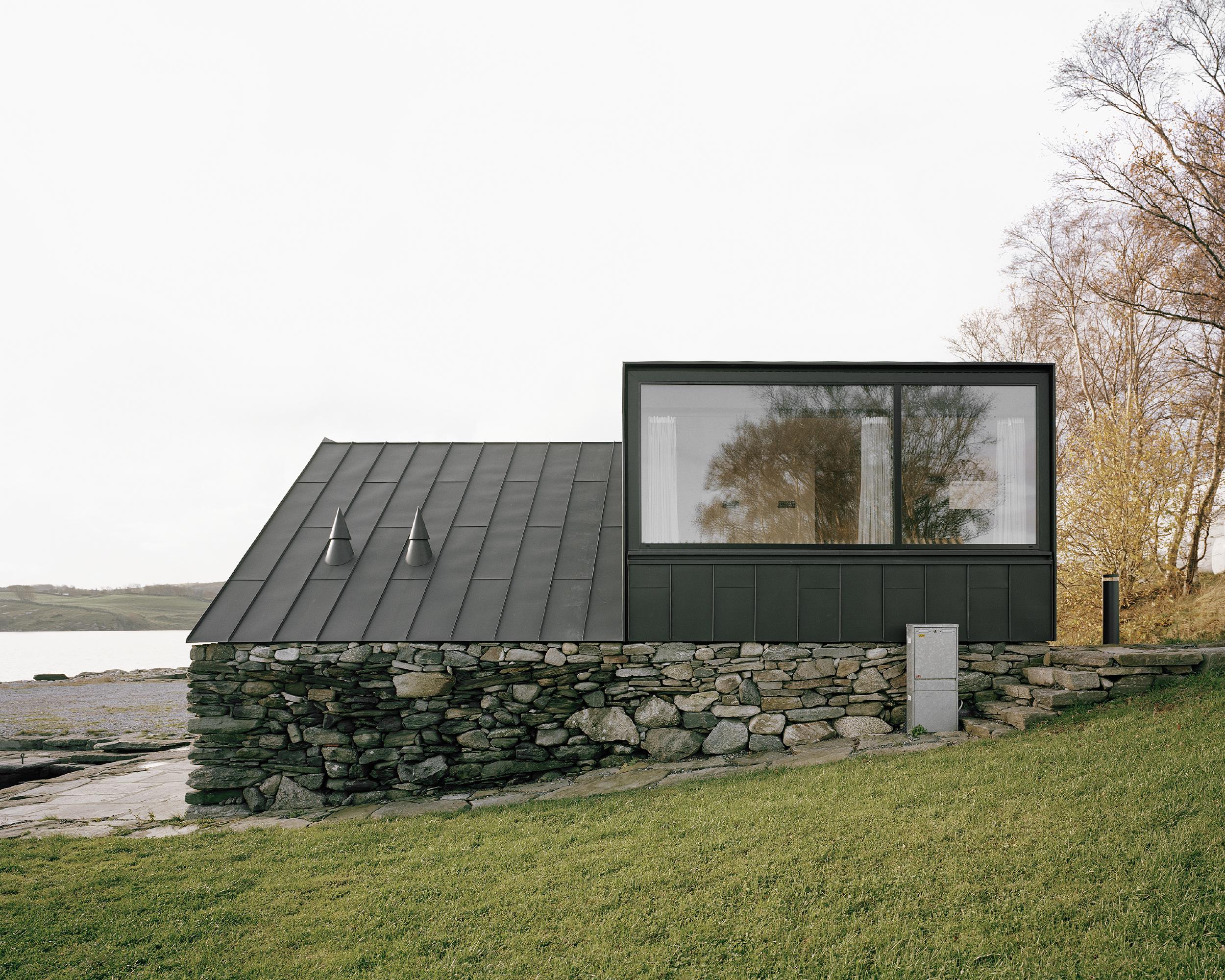 Boat House (Naust) by Espen Surnevik — A modest boathouse sits elevated on a dry-stone foundation, its irregular grey and ochre masonry weathered by coastal exposure.