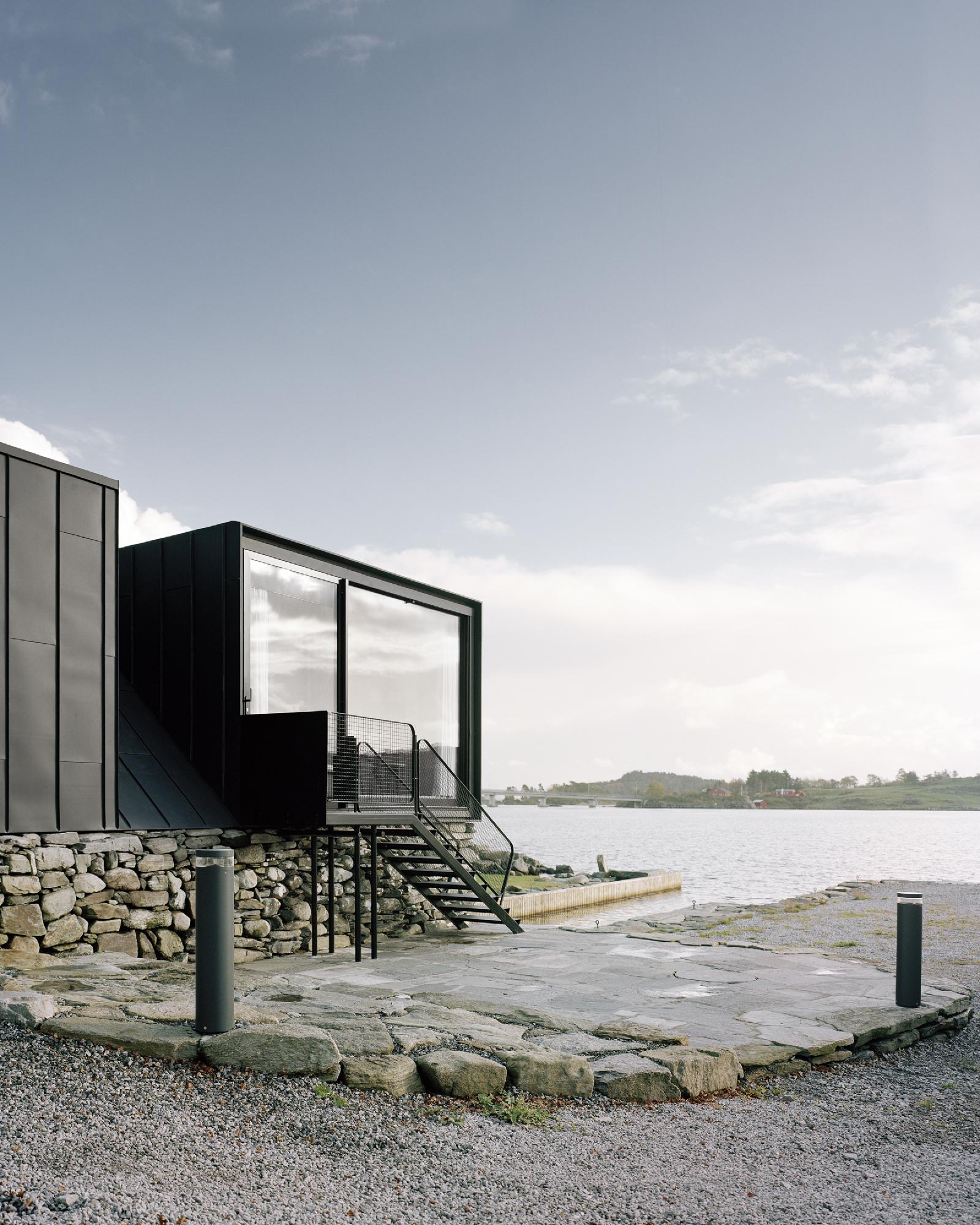 Boat House (Naust) by Espen Surnevik — A black oxidized zinc boathouse elevated on a dry-stone foundation of irregular grey and ochre coursing, positioned at the water's edge in Sokn.