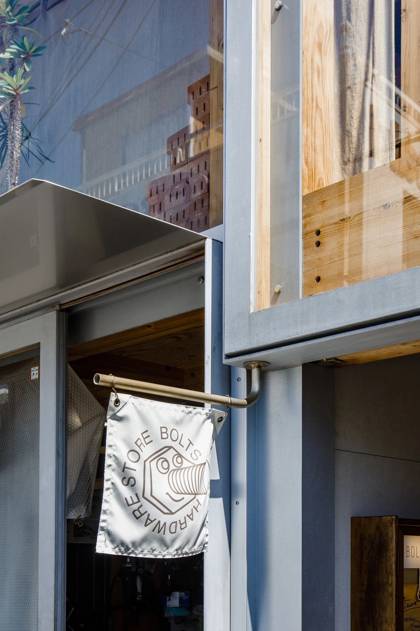 House A/Shop B (Bolts Hardware Store) by KimuraMatsumoto Architects — A storefront banner displaying the Bolts Hardware Store logo hangs from a brass rod mounted to the steel frame of the shop's façade.