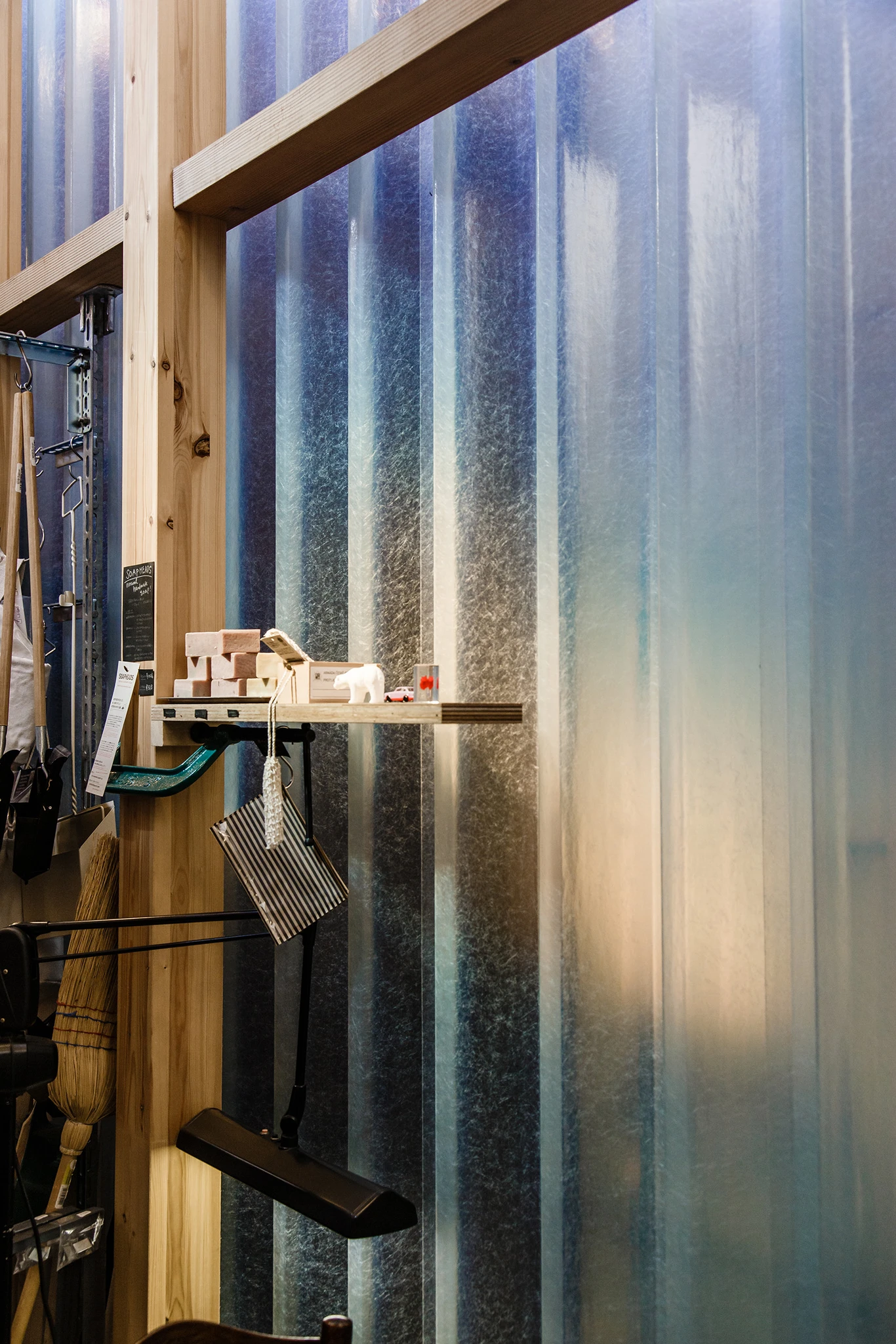 House A/Shop B (Bolts Hardware Store) by KimuraMatsumoto Architects — Interior workshop view through full-height corrugated metal storefront, revealing wooden workbench laden with unfinished wooden blocks and tools.