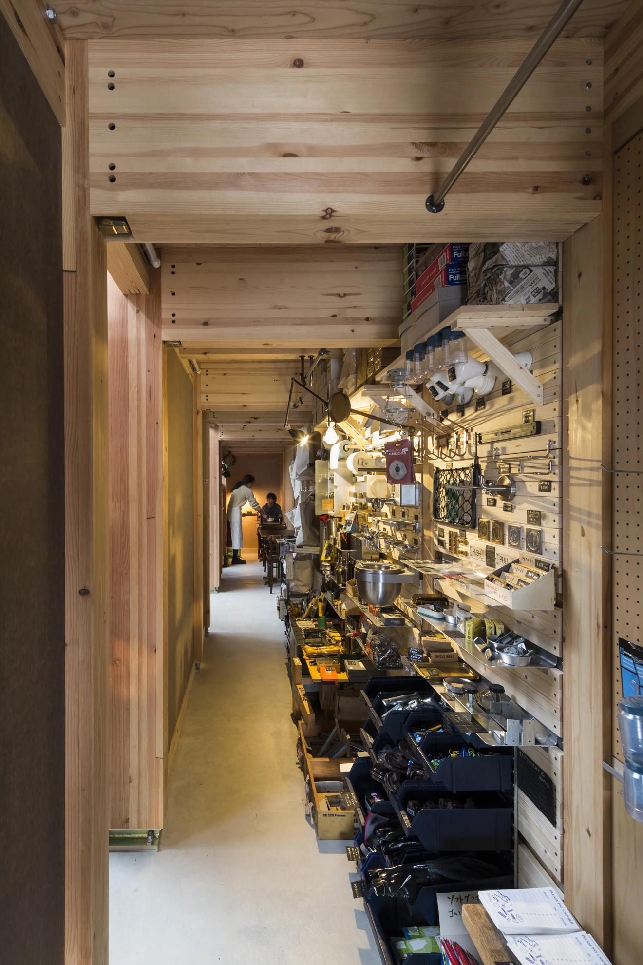 House A/Shop B (Bolts Hardware Store) by KimuraMatsumoto Architects — Interior corridor of a narrow hardware store revealing densely organized merchandise on wall-mounted pegboard and shelving systems.