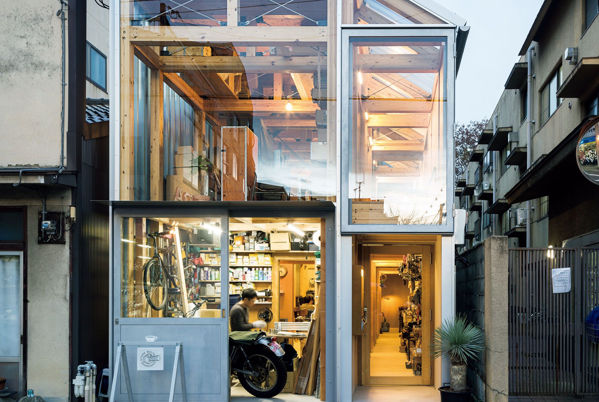 House A/Shop B (Bolts Hardware Store) by KimuraMatsumoto Architects — A narrow timber-framed storefront compressed between concrete residential buildings reveals a bicycle repair workshop at ground level, its interior...