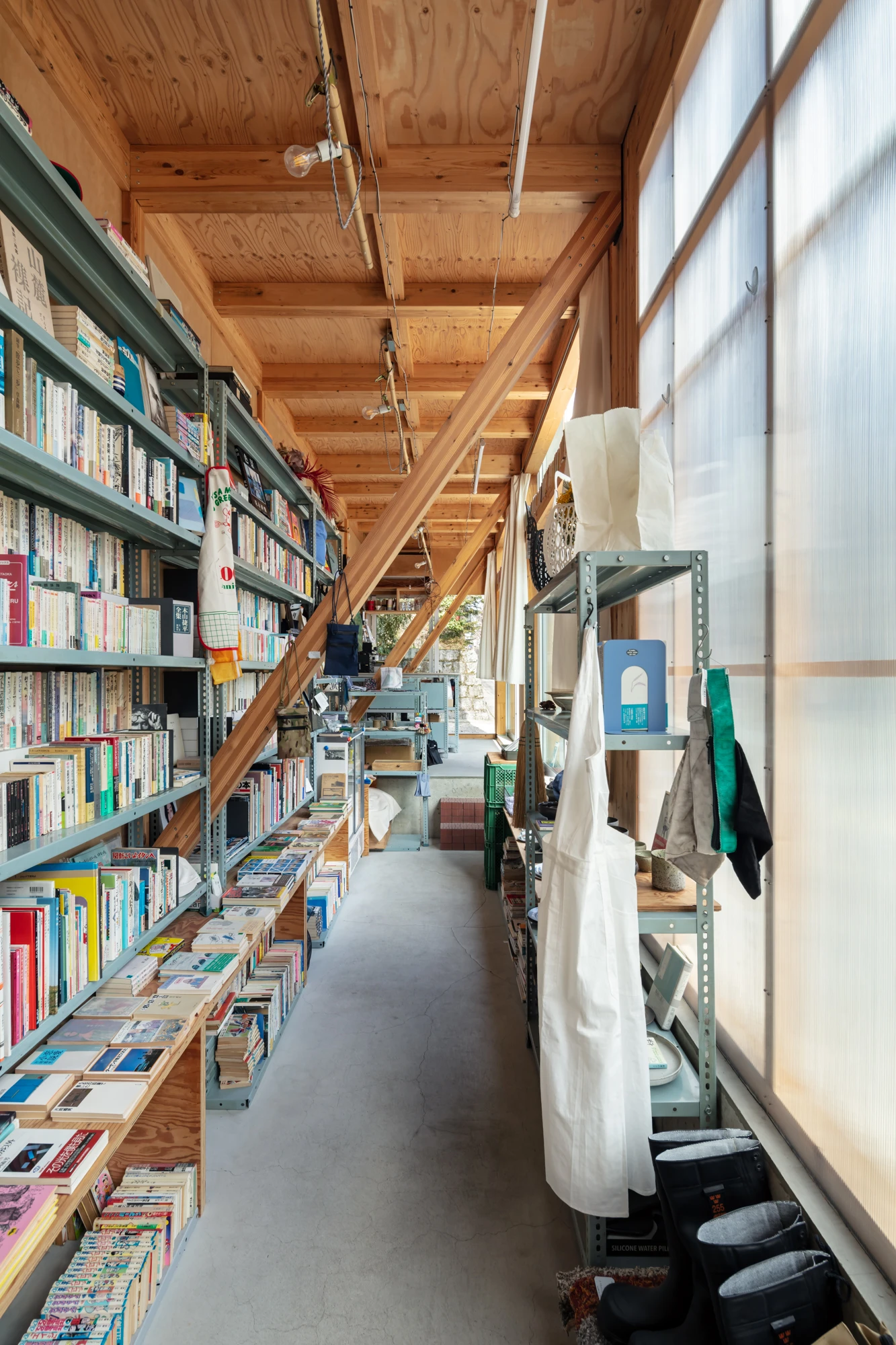 House S/Shop B (ba hütte) by KimuraMatsumoto Architects — Interior view of a narrow commercial corridor lined with dark-framed metal shelving displaying books, DVDs, and media along the left wall.
