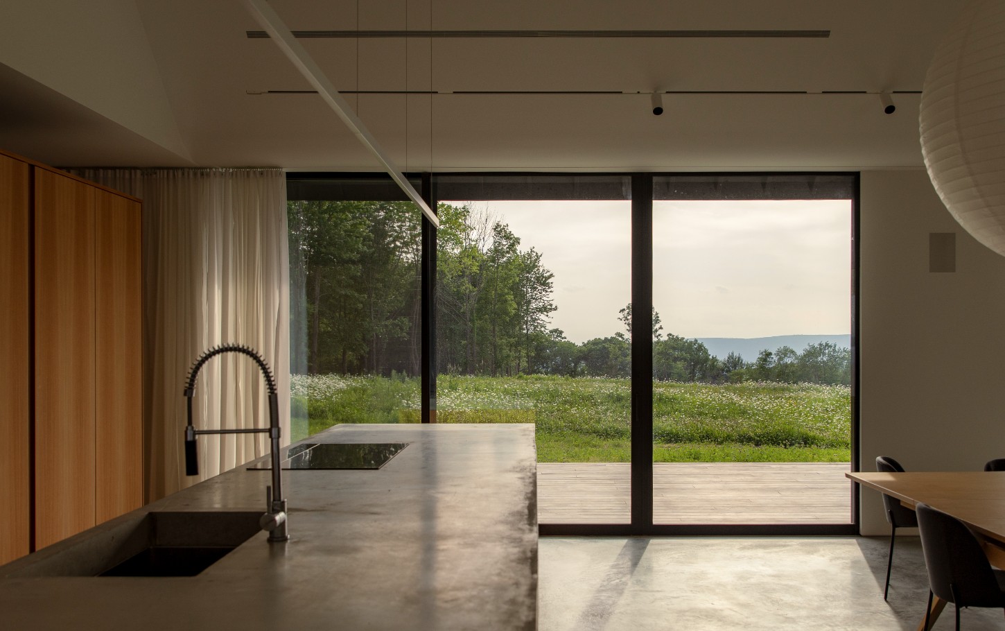 House in a Meadow by Group Projects Architecture — The kitchen interior frames a meadow landscape through floor-to-ceiling glazing, the black corrugated metal cladding visible beyond the glass threshold.