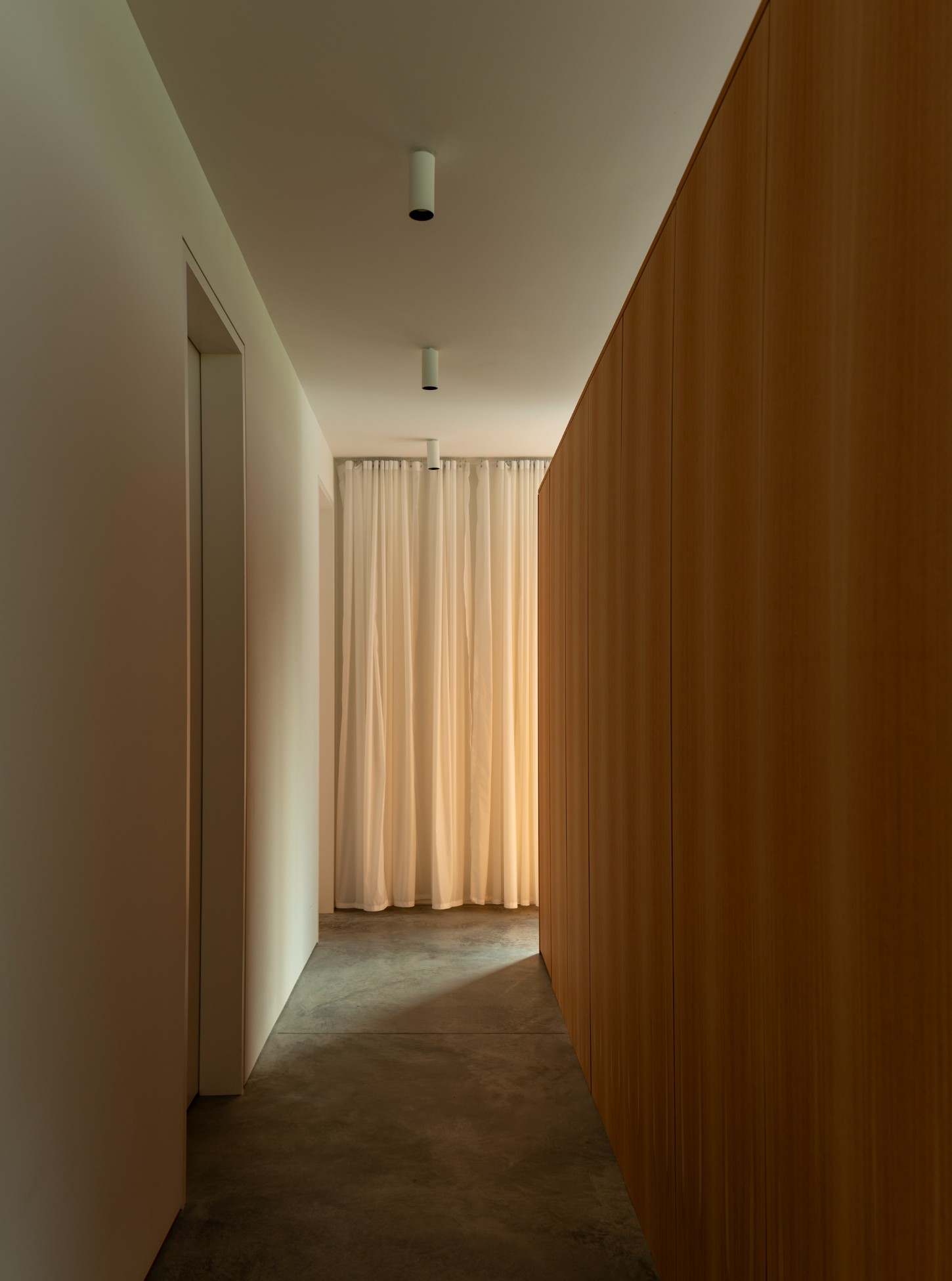 House in a Meadow by Group Projects Architecture — A hallway corridor lined with timber wood paneling on the right wall, white plaster on the left, concrete flooring throughout.