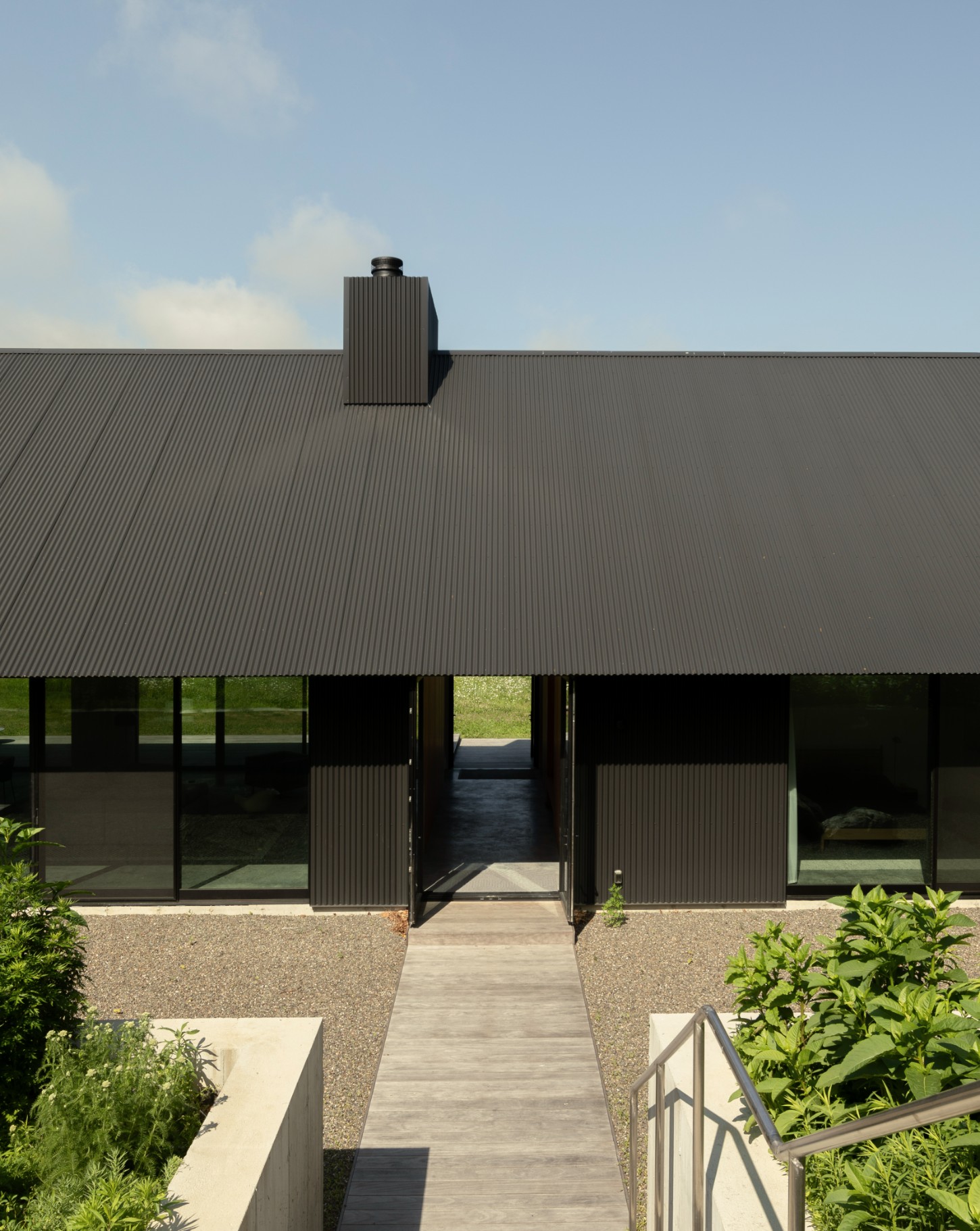 House in a Meadow by Group Projects Architecture — The primary facade of House in a Meadow presents a low-slung horizontal composition dominated by black corrugated metal cladding that extends across the...