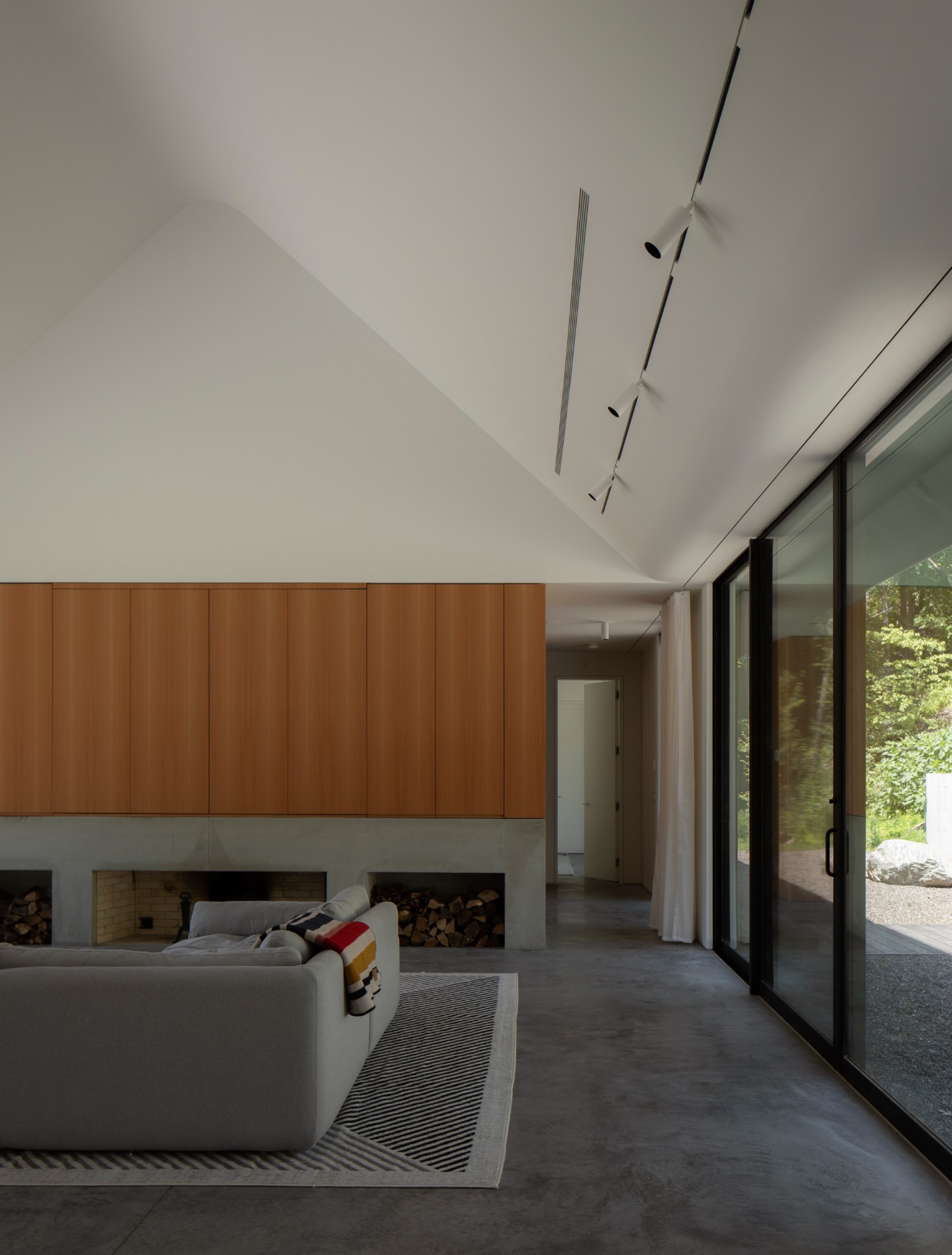 House in a Meadow by Group Projects Architecture — Interior view of the living space revealing the wood-clad feature wall in warm timber tones, with twin firewood storage alcoves set into a concrete plinth.