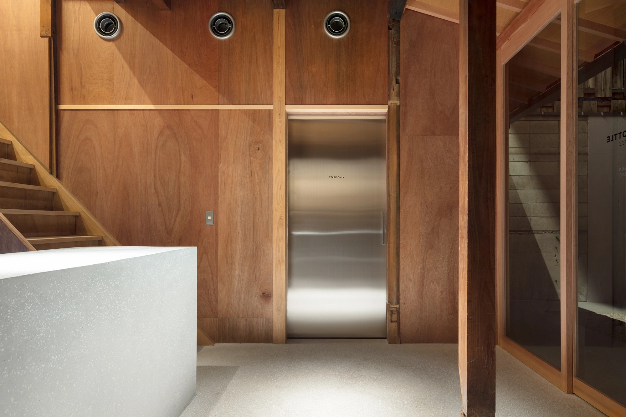 Blue Bottle Coffee Kyoto Cafe by Schemata Architects — A staff-only elevator finished in brushed stainless steel occupies the central passage between timber-clad walls, its reflective surface contrasting sharply...