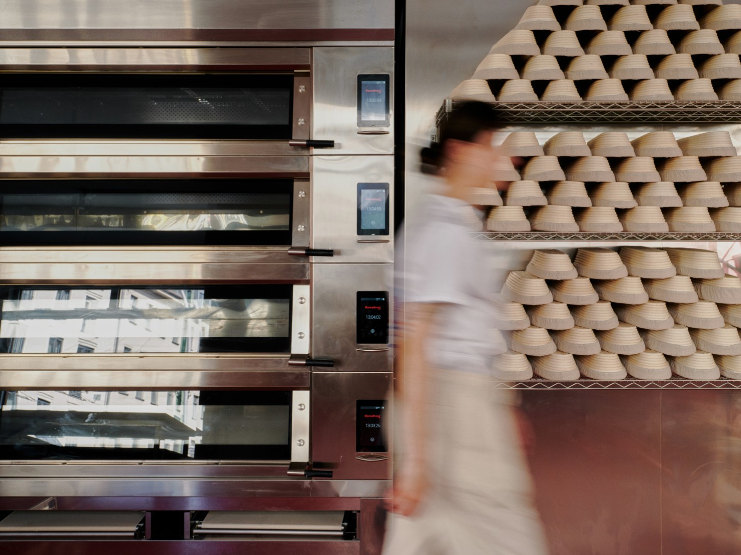 ACID Cafe by Plantea Studio by Plantea Estudio — A motion-blurred figure moves through the ACID Cafe's production zone, captured mid-stride past tiered wooden bread baskets and a vertical stack of...