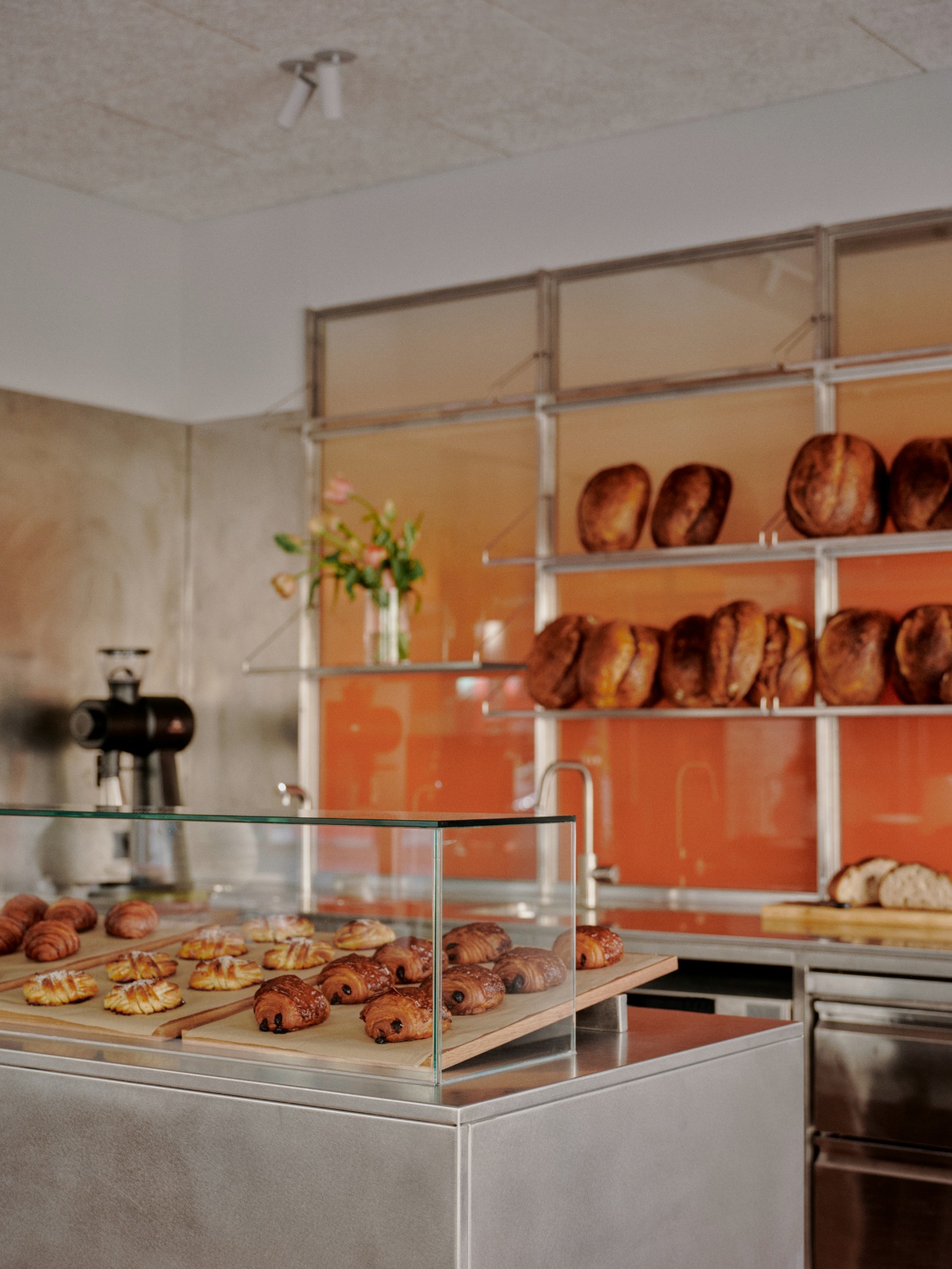 ACID Cafe by Plantea Studio by Plantea Estudio — The display counter anchors the foreground with croissants and pain au chocolat arranged on wooden boards beneath protective glass, while vertical...