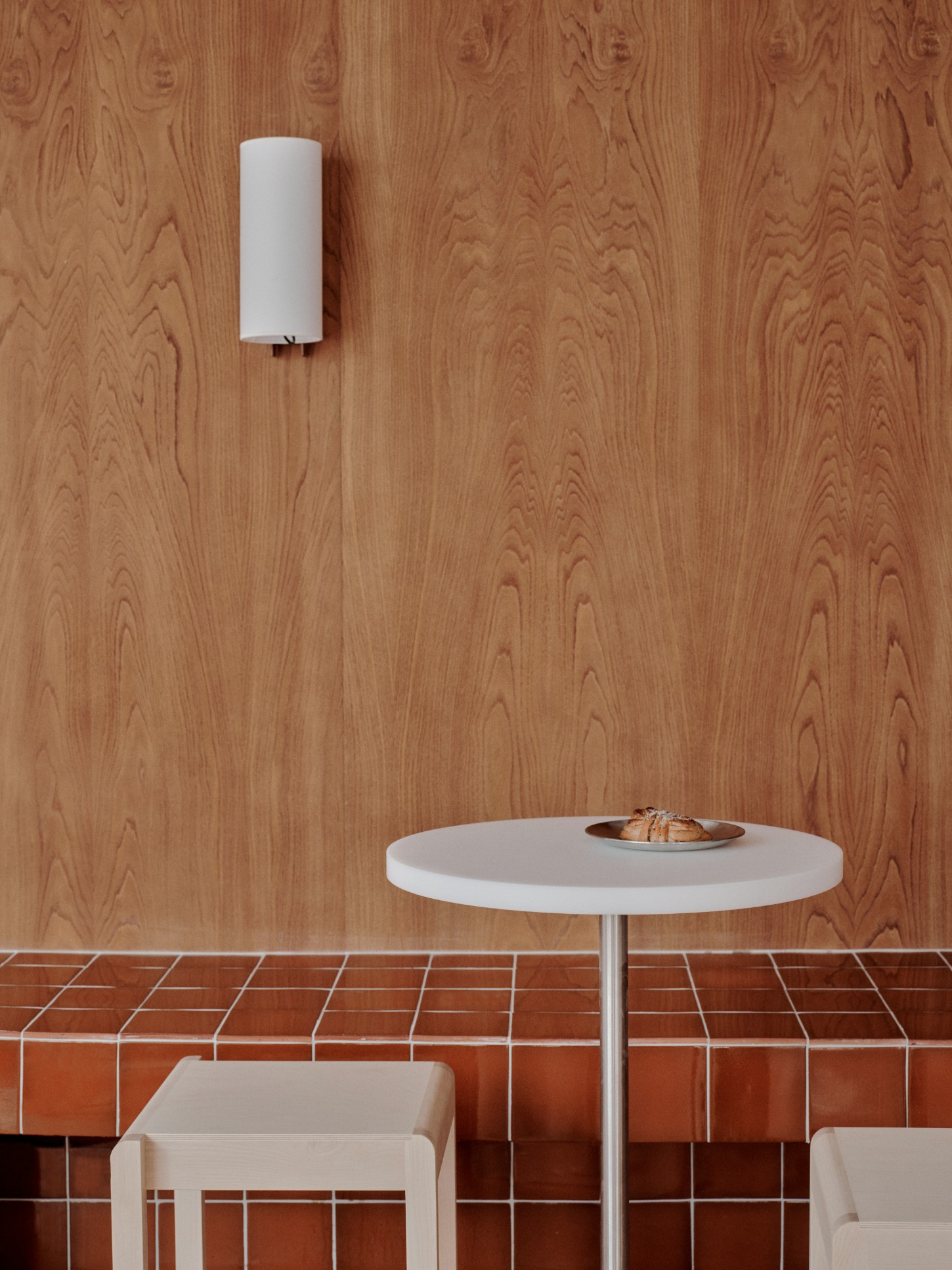 ACID Cafe by Plantea Studio by Plantea Estudio — A minimalist café vignette centered on a white circular table with polished metal pedestal base, positioned against a warm timber wall with pronounced...