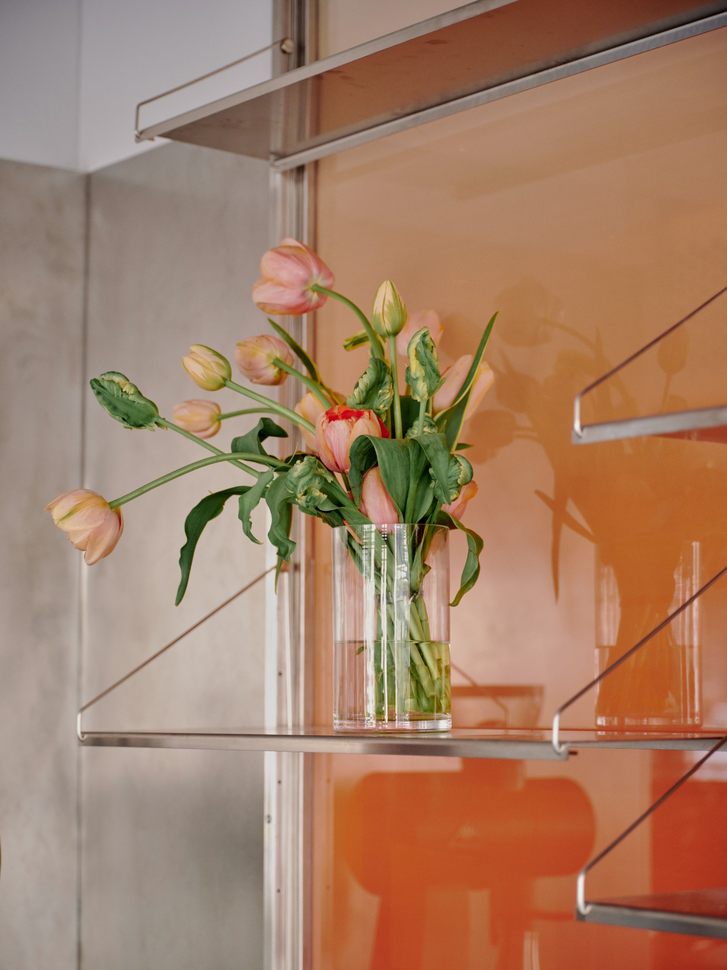 ACID Cafe by Plantea Studio by Plantea Estudio — A clear glass vase containing pink and cream tulips at various stages of bloom sits on a metal-framed shelf against an orange-toned glazed wall panel.