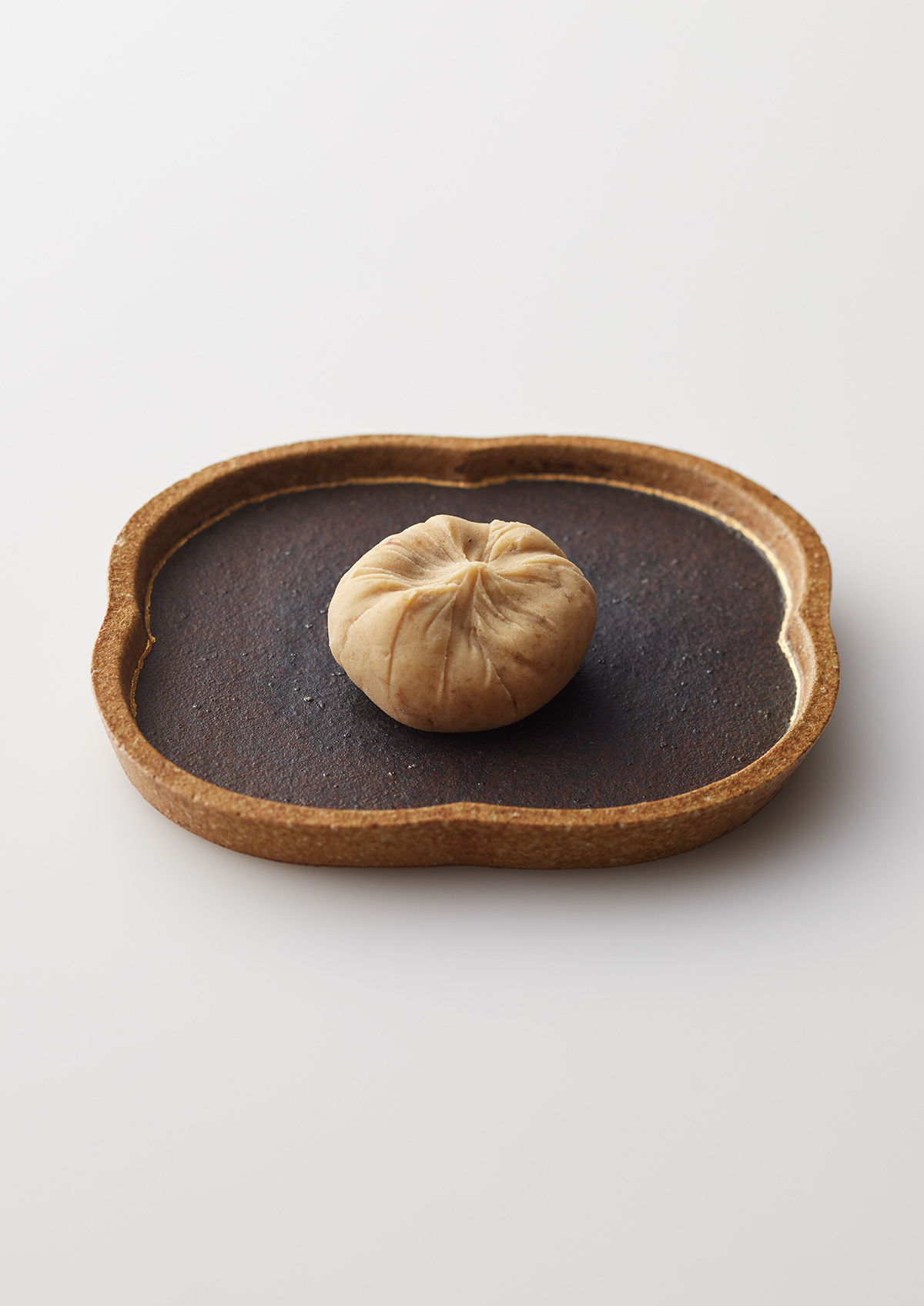 La Collina Ōmi-Hachiman by Terunobu Fujimori + Michele De Lucchi — A minimalist tabletop composition featuring a single pleated dumpling resting on a dark slate surface within a rounded rectangular earthen vessel.