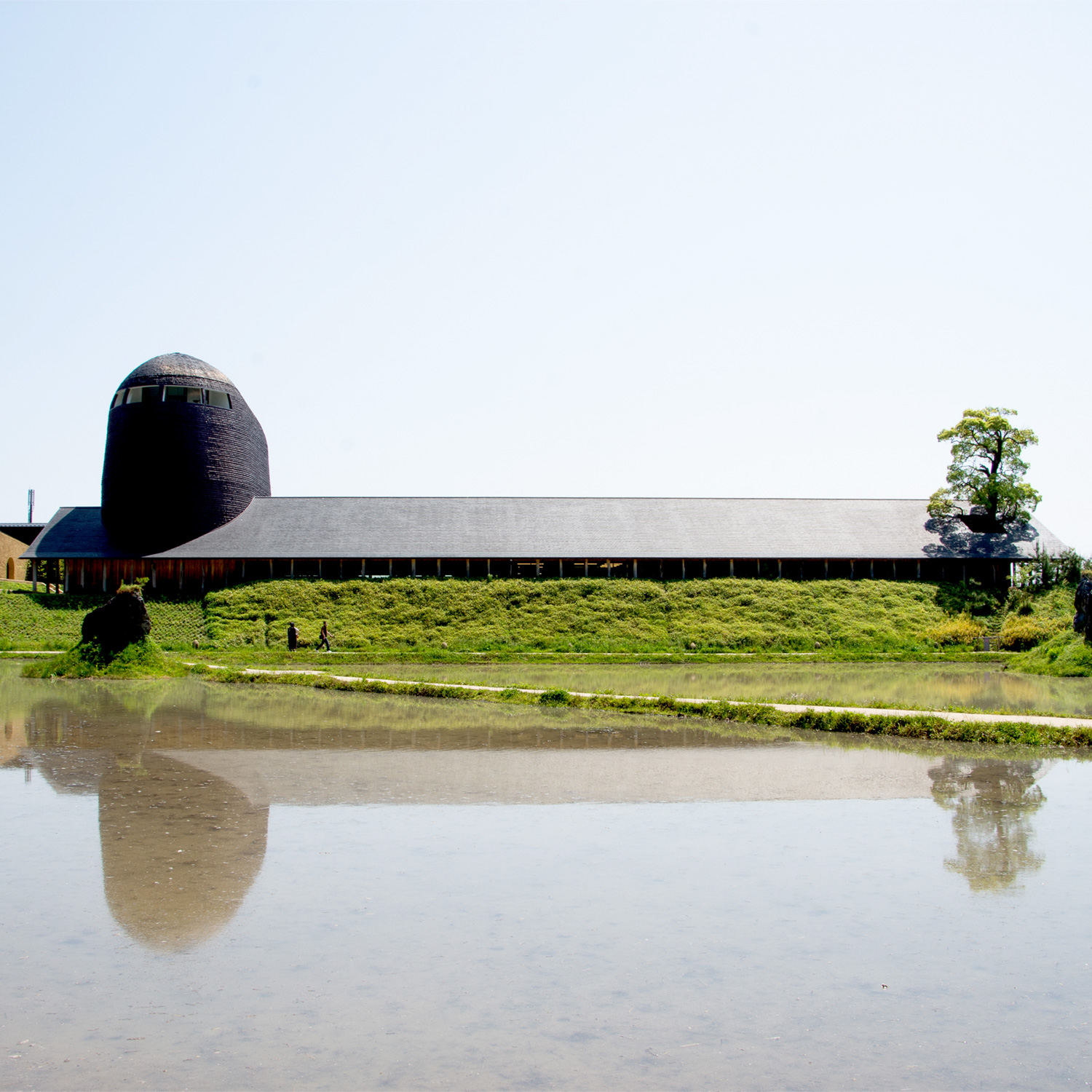 La Collina Ōmi-Hachiman by Terunobu Fujimori + Michele De Lucchi — La Collina's vernacular-scaled timber barn extends horizontally across the frame, its dark pitched roof anchoring a composition dominated by dense green...