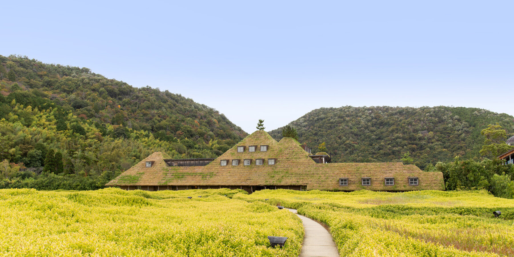 La Collina Ōmi-Hachiman by Terunobu Fujimori + Michele De Lucchi — A monolithic brick structure with a pitched roof sits anchored within a cultivated landscape of bright yellow-green vegetation.
