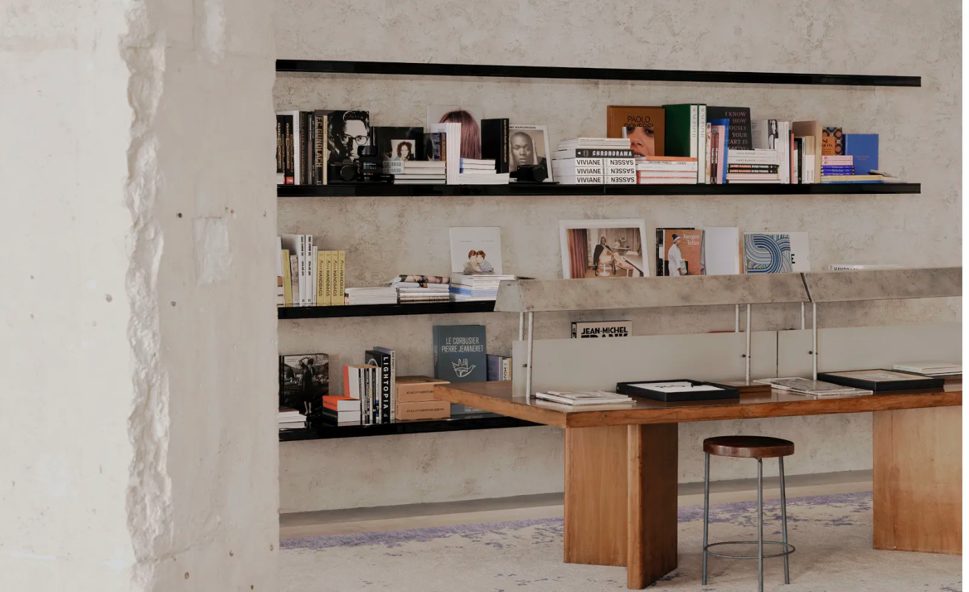 Saint Laurent Babylone in Paris — The workspace anchors itself around a curved timber table with organic proportions, paired with metal bar stools featuring thin angular legs that create...