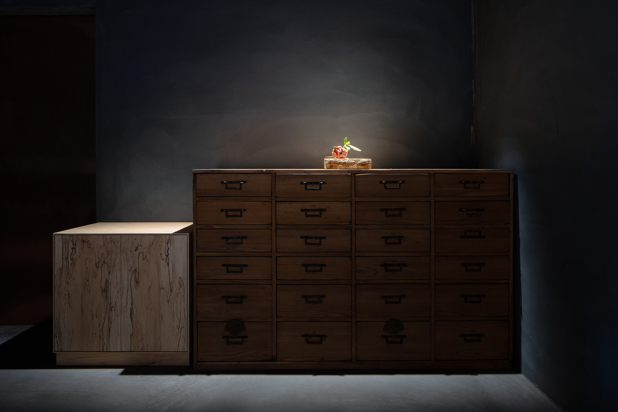 mitomi by Koyori + Aterier Salt — A wooden storage unit occupies the foreground of this darkened interior, its light-toned timber surface with visible grain pattern contrasting sharply...