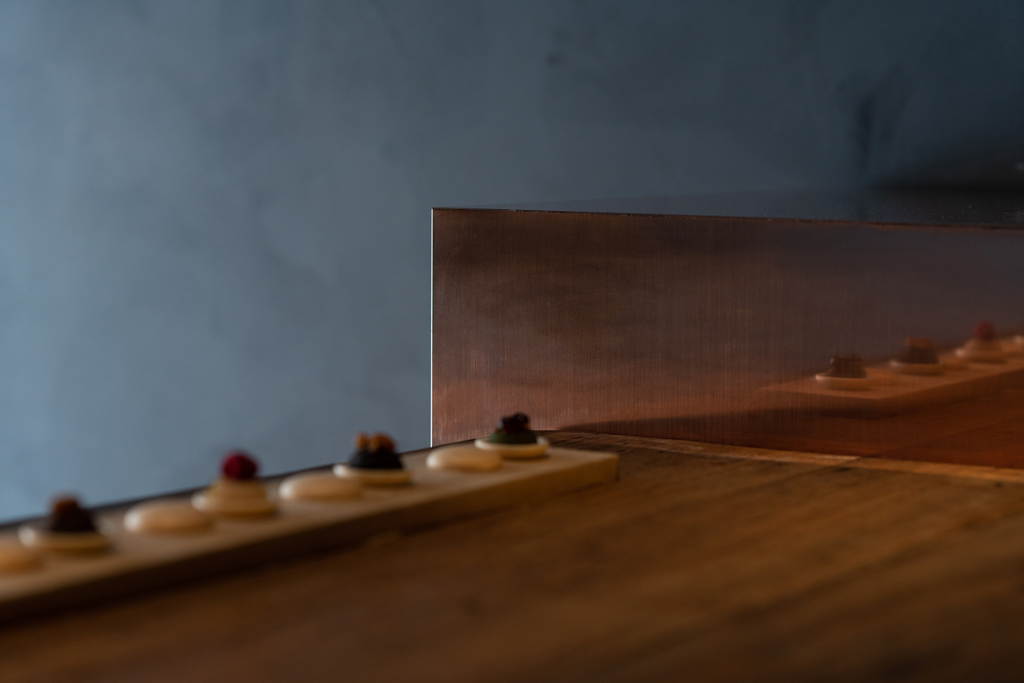 mitomi by Koyori + Aterier Salt — A linear display arrangement recedes toward a copper-clad vertical panel, with small monaka sweets positioned along a timber shelf.