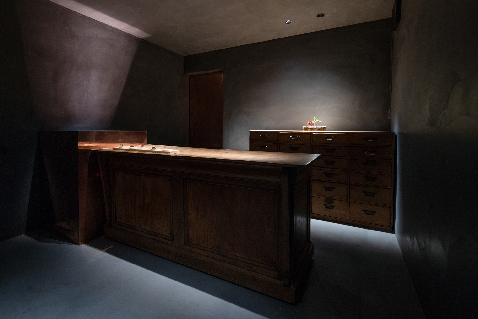 mitomi by Koyori + Aterier Salt — A wooden counter occupies the foreground of this Kyoto tea-inspired shop interior, its light timber surface elevated against deep charcoal render covering...