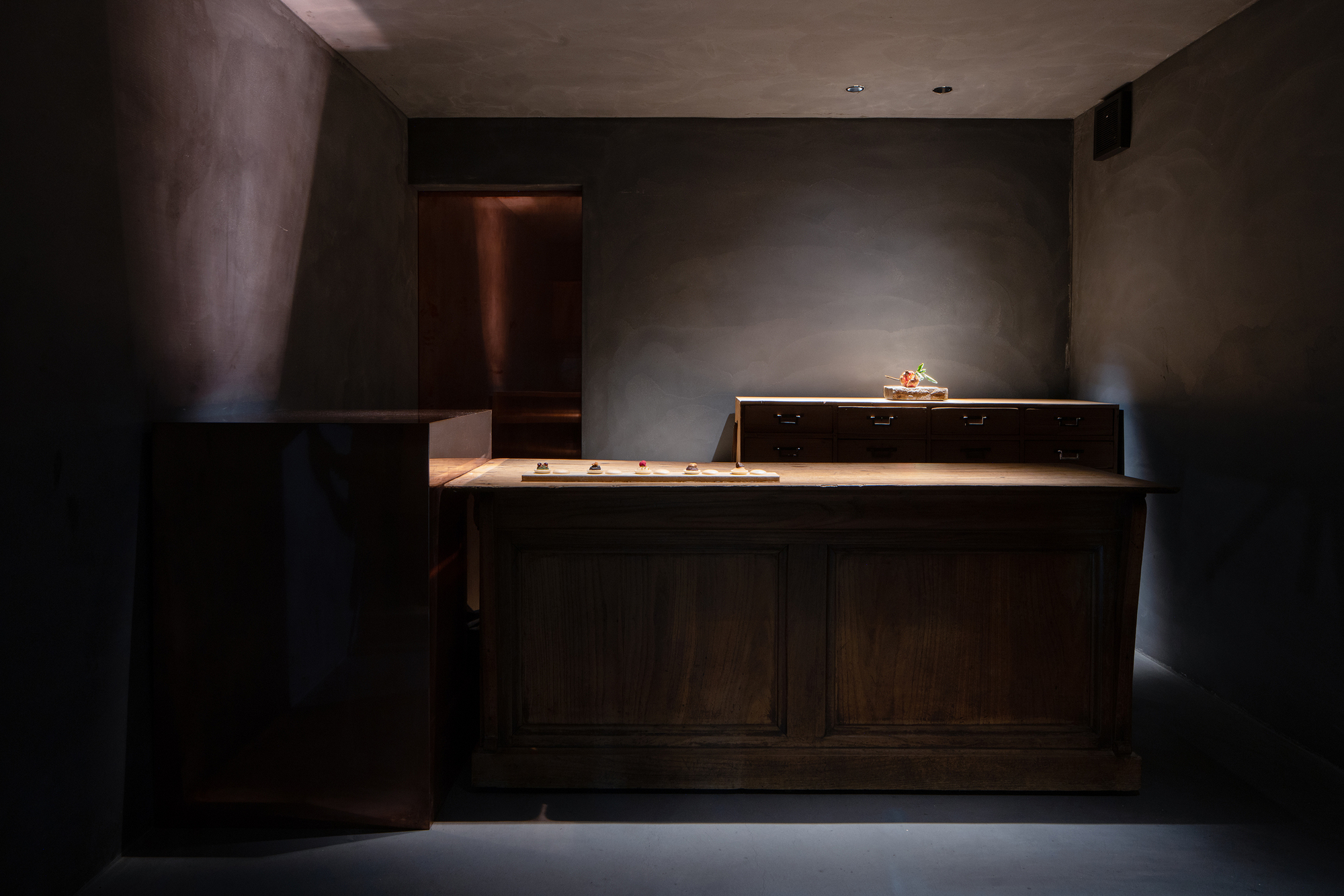 mitomi by Koyori + Aterier Salt — A wooden display counter extends horizontally across a darkened interior, its surface illuminated by a concentrated overhead beam that carves through the...