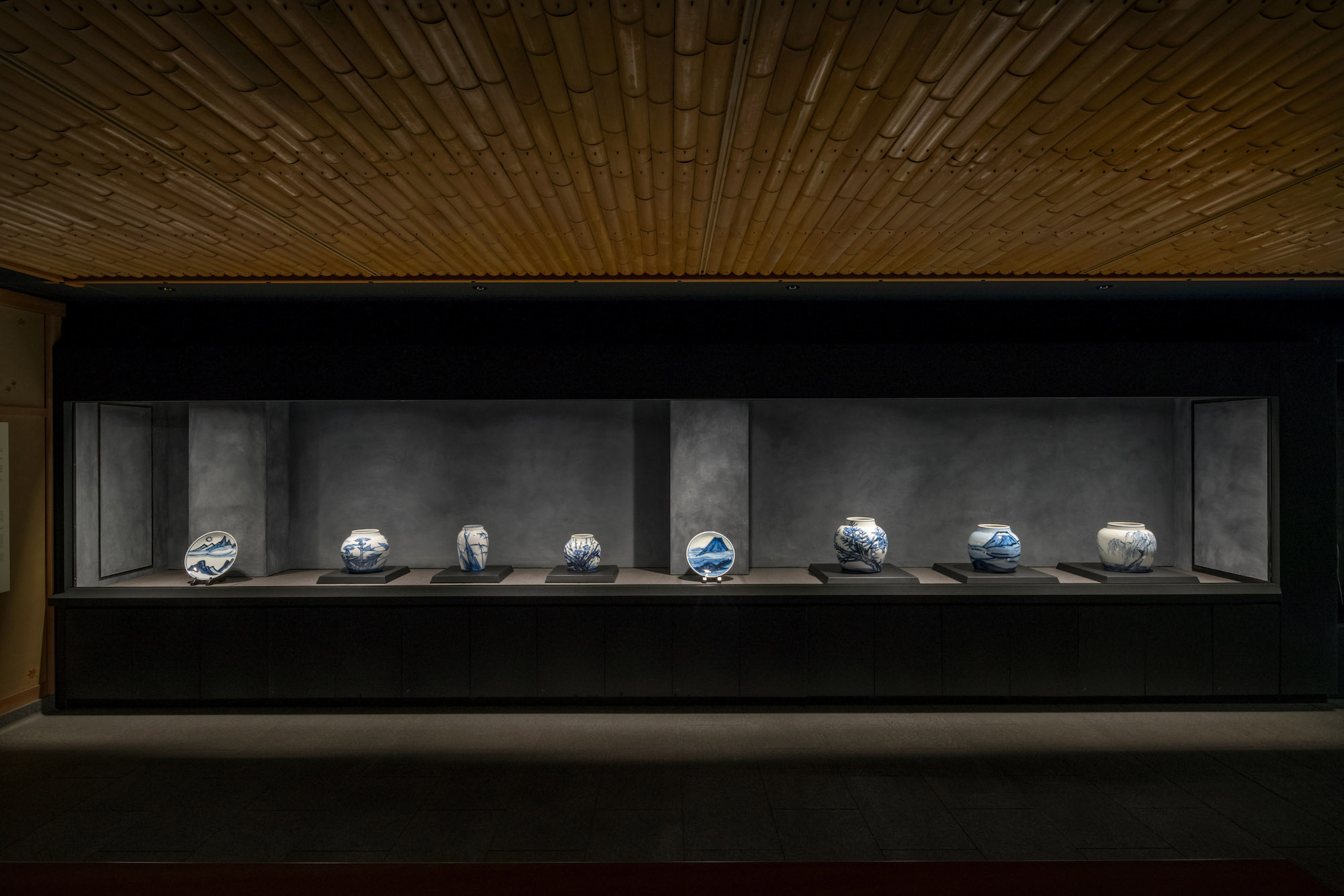 Kondo Museum in Kyoto by Mamiya Shinichi Design Studio — A linear display case stretches across the gallery wall, housing eight blue-and-white ceramic vessels arranged in regular intervals on a dark shelf.