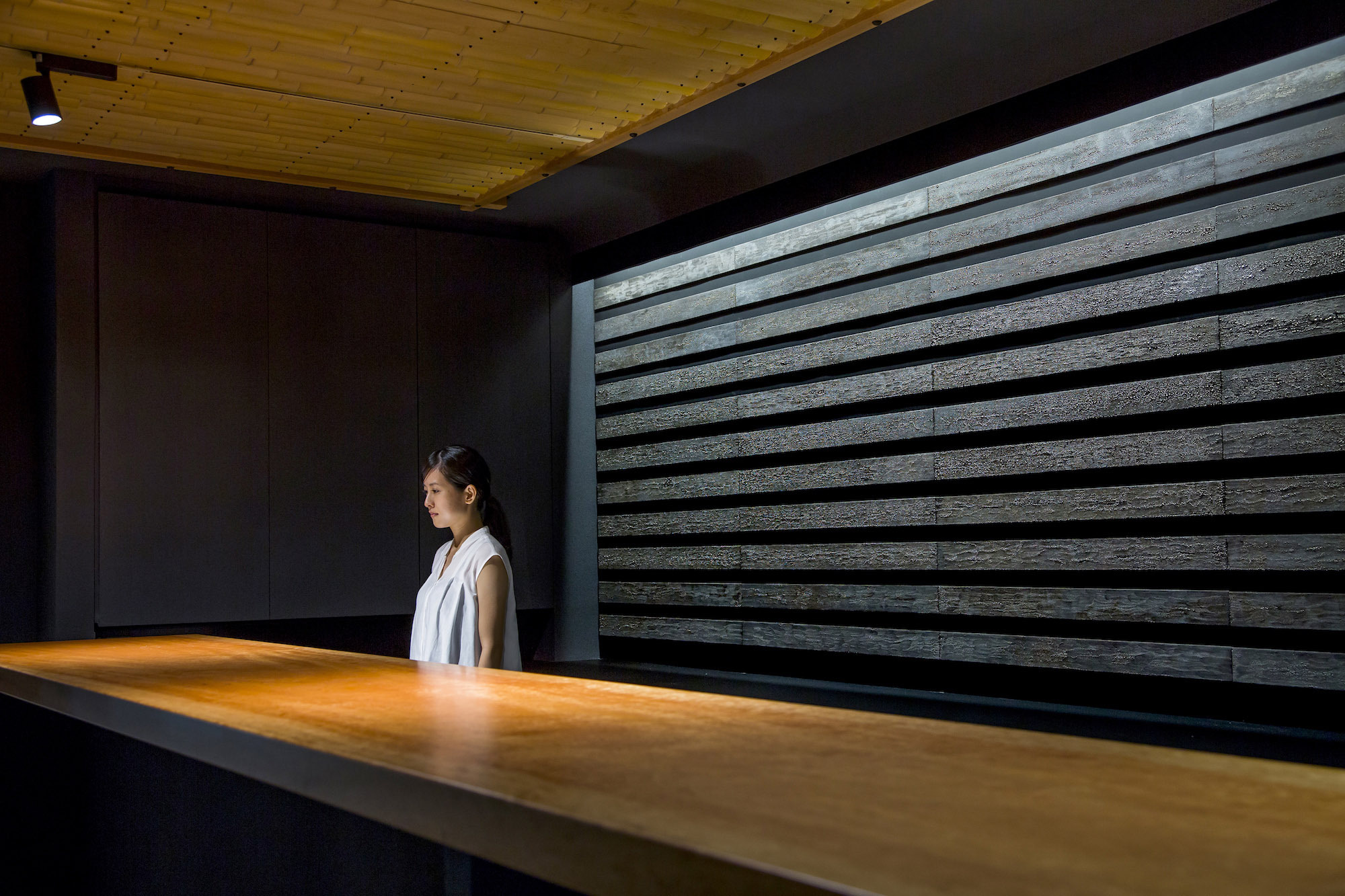 Kondo Museum in Kyoto by Mamiya Shinichi Design Studio — A woman in a white sleeveless vest stands in profile within the museum's gallery space, positioned before a horizontal timber lattice wall with regular...