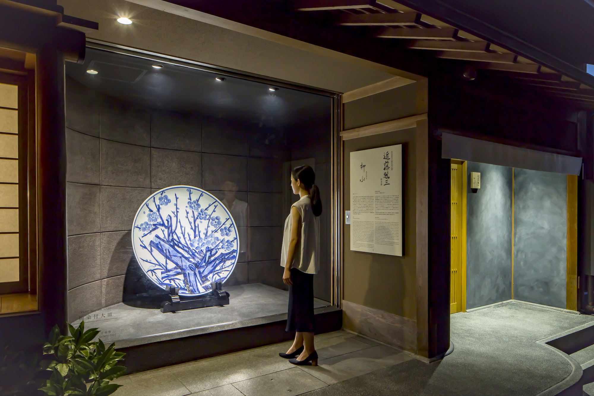 Kondo Museum in Kyoto by Mamiya Shinichi Design Studio — A visitor stands before an illuminated ceramic plate featuring blue-and-white decoration, recessed within a darkened gallery niche lined with charcoal-toned...