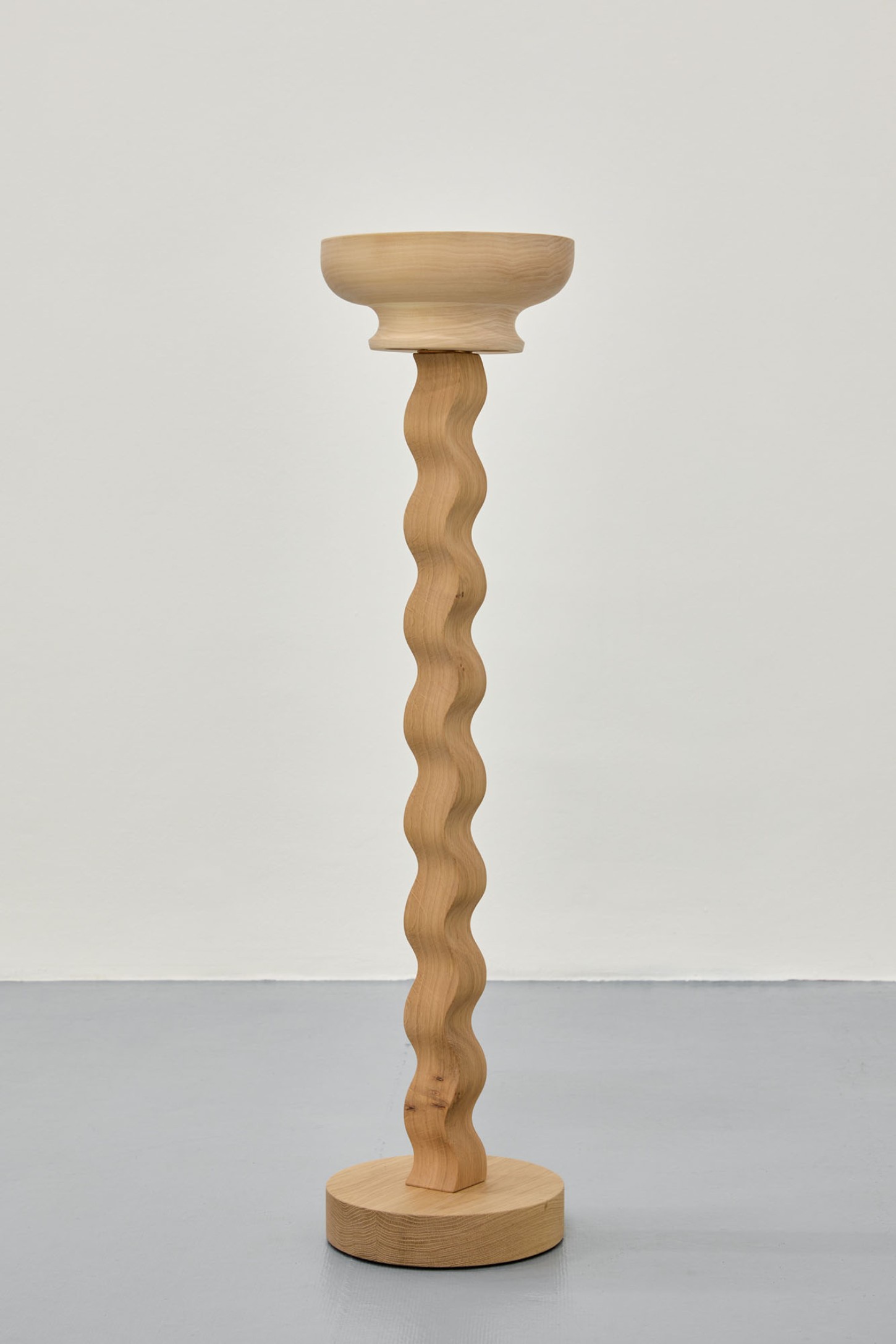 Thirst by Eliška Konečná at Polansky — A tall wooden pedestal carved from light oak with a sinuous, undulating shaft that spirals from a circular base to a wide, shallow bowl-form top.