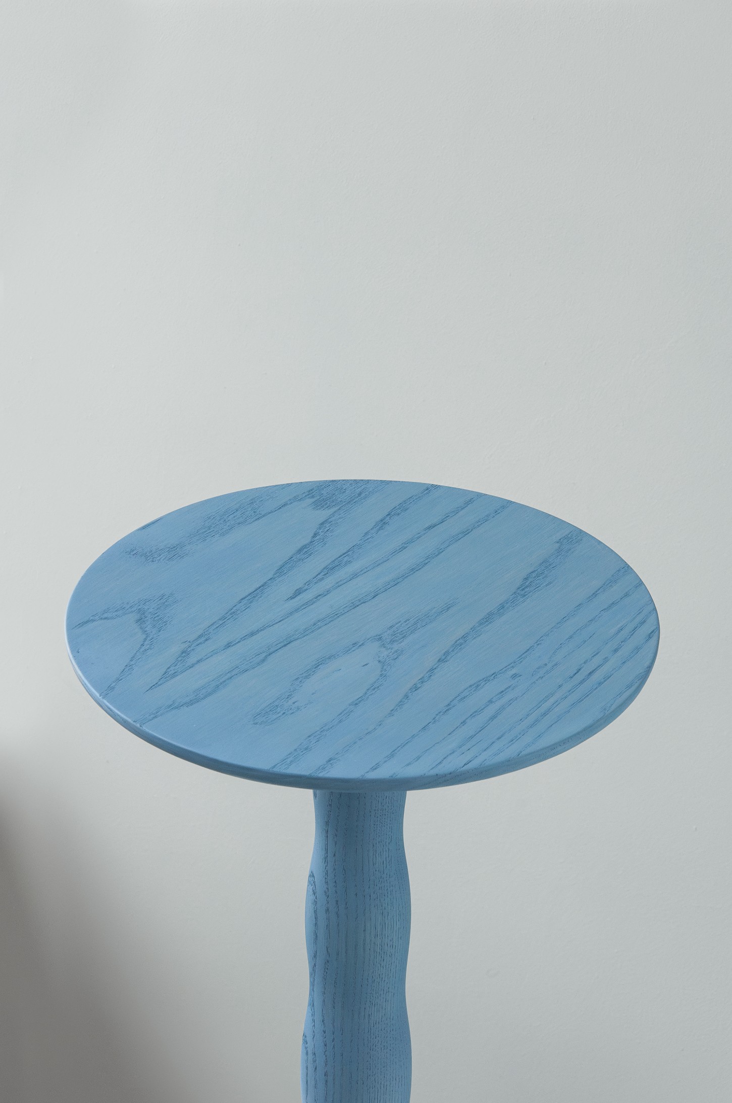 'Burn & Turn' by Adrian Bursell + Siri Svedborg — A turned timber pedestal table with pale periwinkle finish, the cylindrical column displaying pronounced vertical fluting that creates linear shadow patterns.