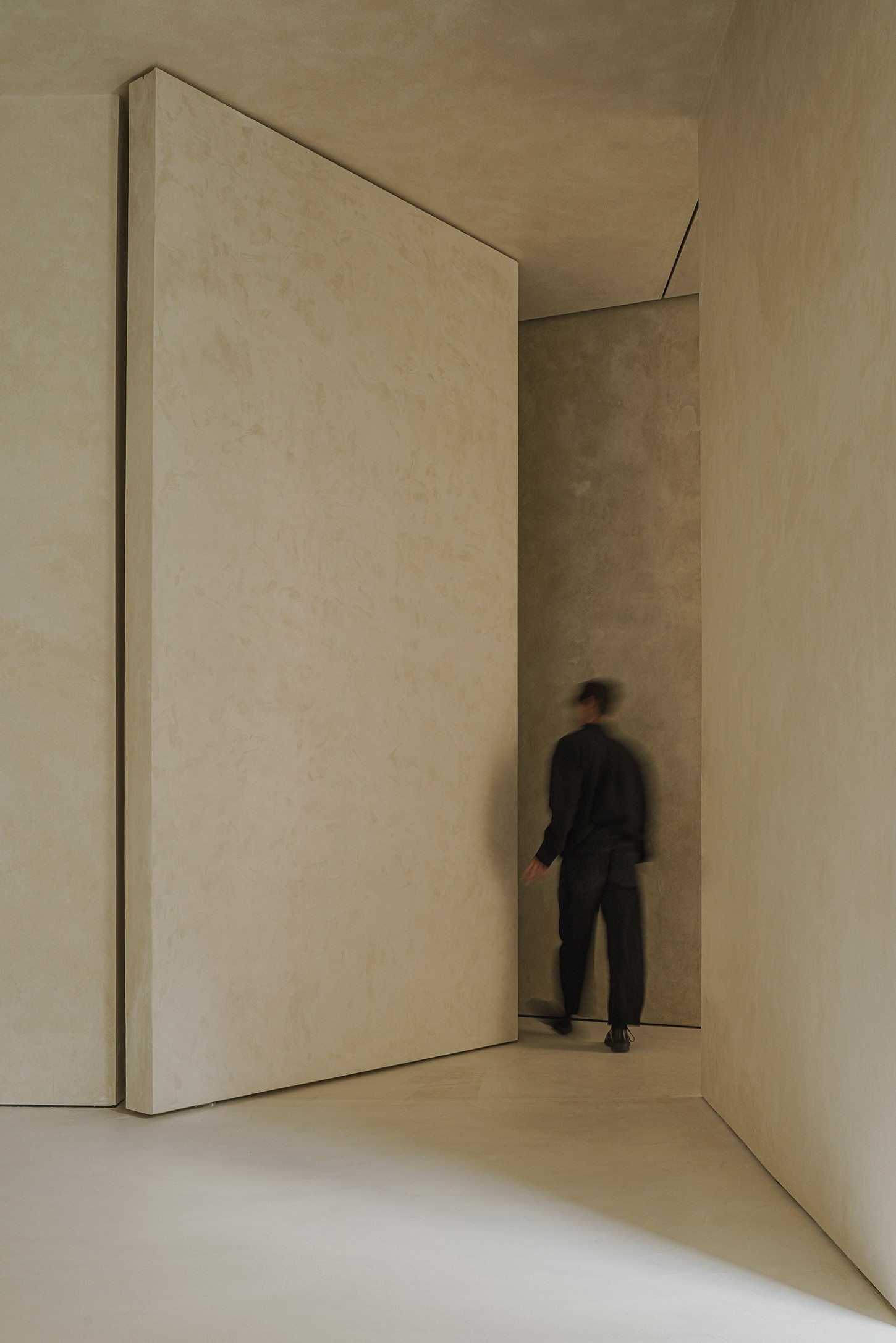 ZØ Residence by Studio Beta Ø — A figure in dark business attire stands within a concrete passage, framed by a monumental cream-colored plaster panel that pivots open like a door.