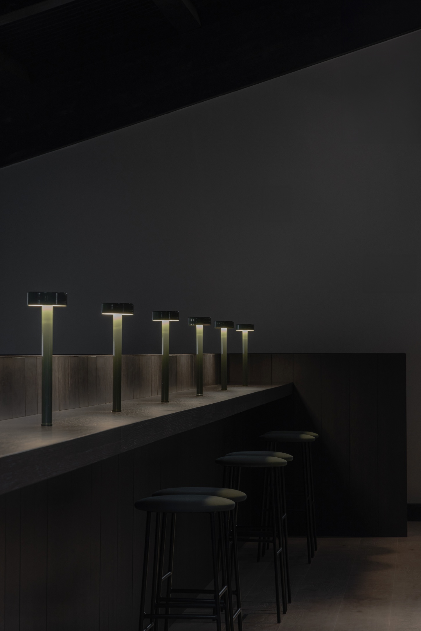 Tritium Winery by Francesc Rifé Studio — A dimly lit bar counter features a receding line of six illuminated T-shaped fixtures with brass tops mounted on a wood-paneled wall, their light casting a...