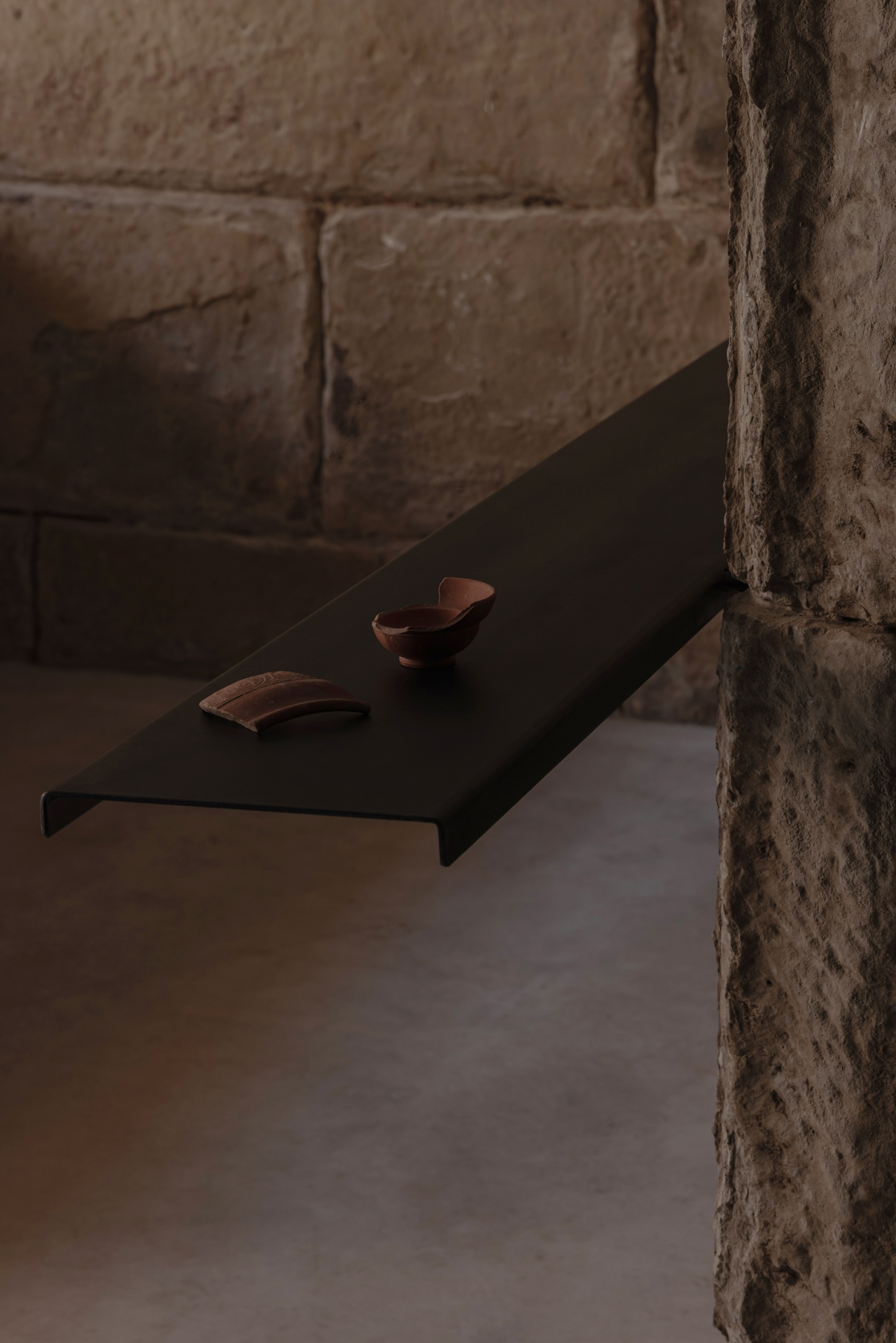 Tritium Winery by Francesc Rifé Studio — A black-painted timber shelf mounted to a weathered stone wall displays two terracotta vessels in varying states of fragmentation.