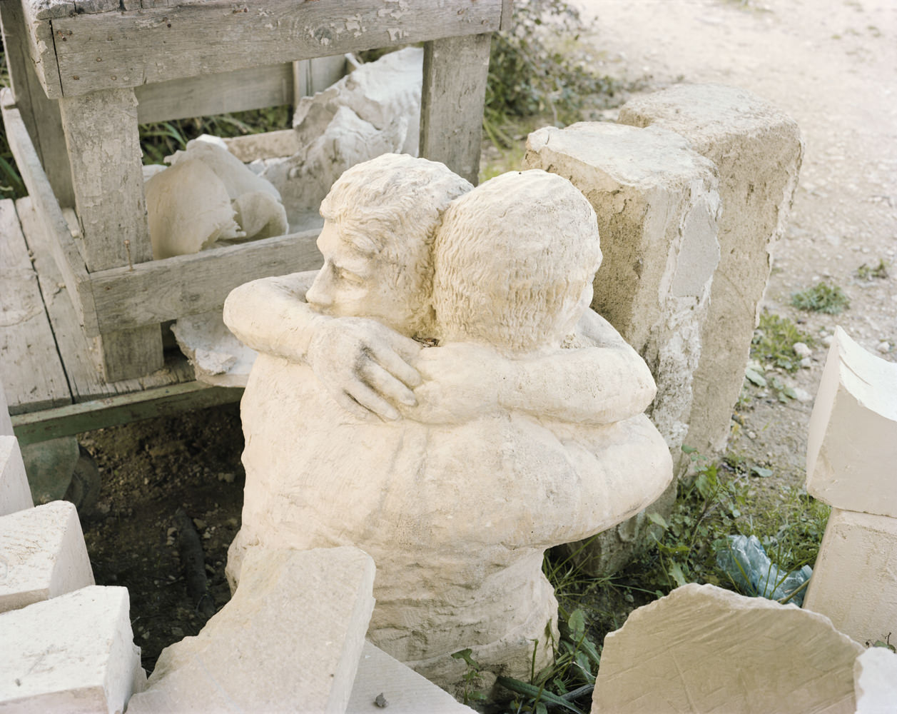 Ghosts in the Field by Murray Ballard — A limestone bust depicting a male figure with curled hair and defined facial features sits among weathered stone blocks and wooden structural elements.
