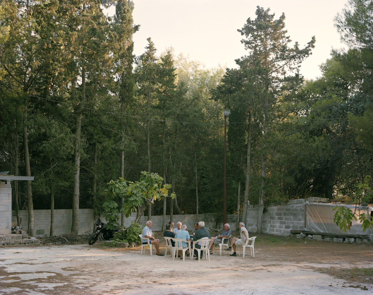 Ghosts in the Field by Murray Ballard — A group of seven elderly residents sit in white plastic chairs arranged in a circle on unpaved ground, engaged in conversation within a walled compound.