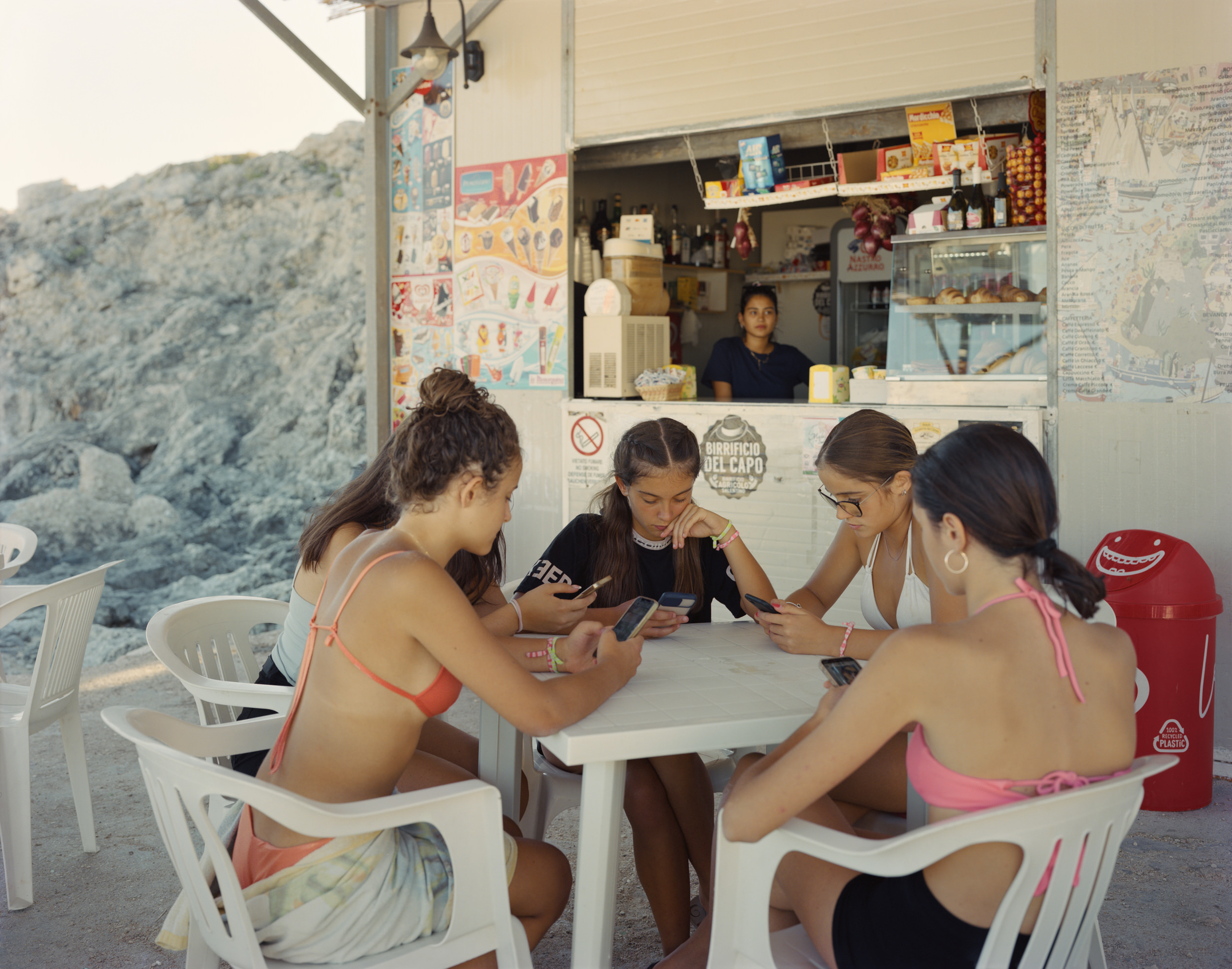 Ghosts in the Field by Murray Ballard — Four young women seated around a white plastic table at a beach kiosk in Puglia, each holding mobile phones.