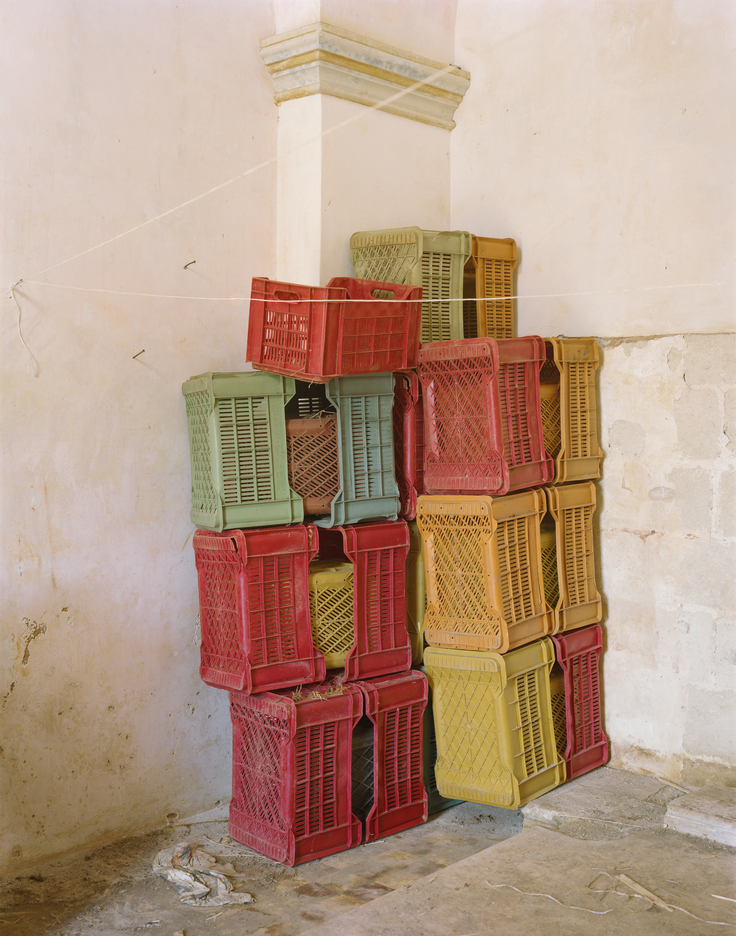Ghosts in the Field by Murray Ballard — Stacked plastic crates in red, yellow, and sage green occupy a corner of a whitewashed interior, their ventilated surfaces creating a geometric tower...