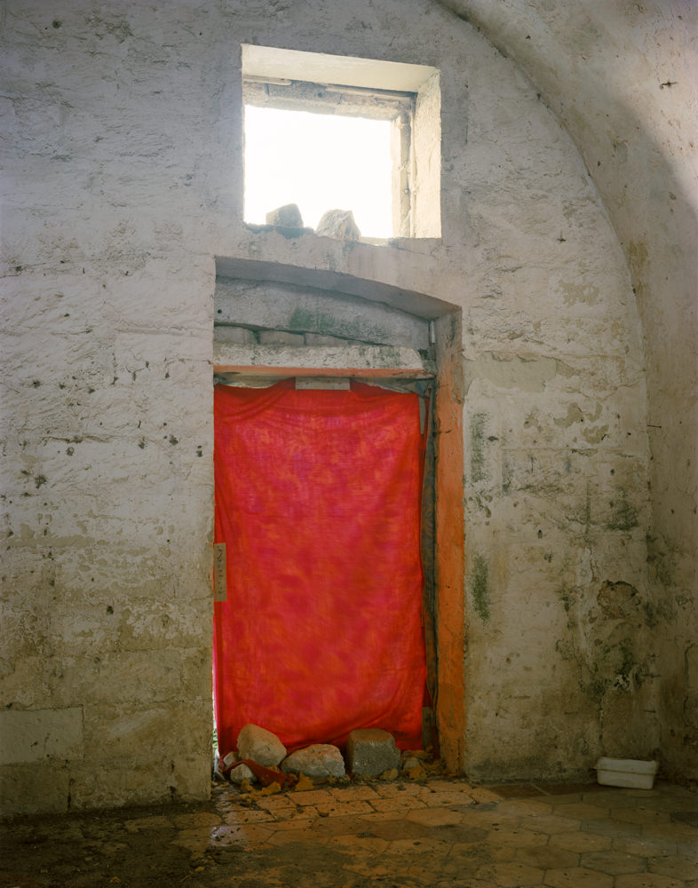 Ghosts in the Field by Murray Ballard — A doorway in an abandoned structure reveals a red fabric suspended across its frame, blocking passage into darkness beyond.