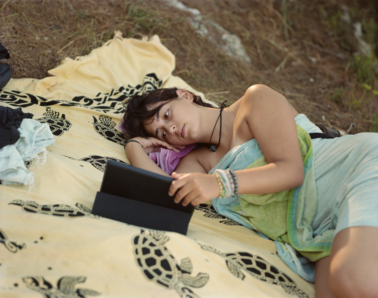 Ghosts in the Field by Murray Ballard — A young person reclines on a patterned fabric spread across sandy ground, holding a tablet device.