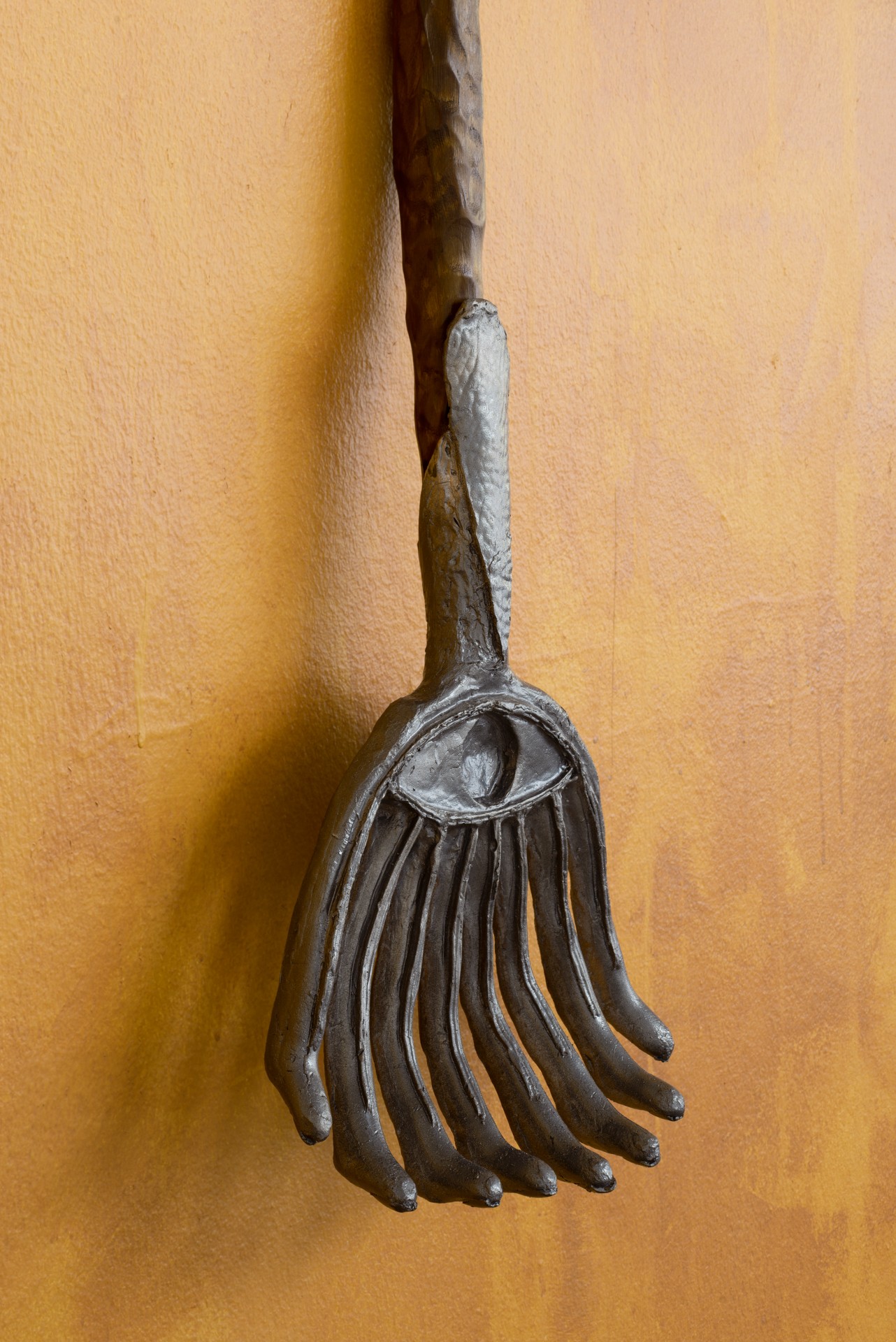 Bleak Harvest by Anna Bak at Vestjyllands Kunstpavillon — A cast-bronze hand suspended from a wooden dowel against an ochre-rendered wall.
