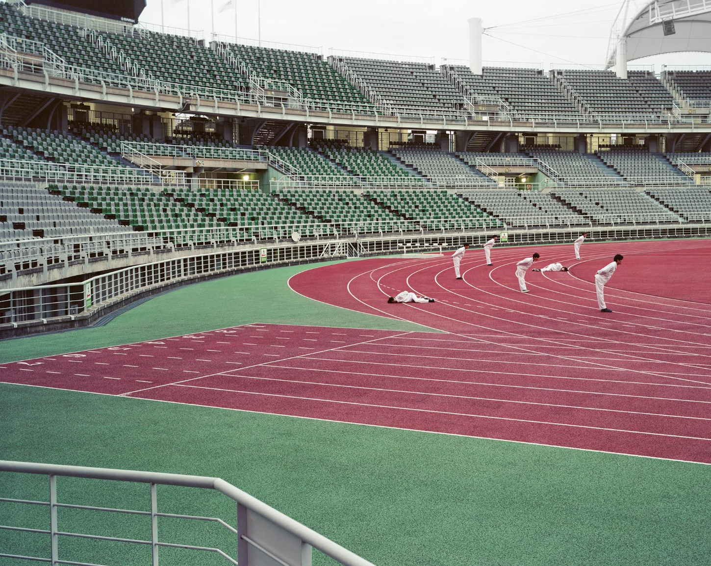 "Wandering, Wondering' by Seok-Woo Song — Five figures in white uniforms positioned across a burgundy running track within an oversized Olympic stadium, their bodies arranged at deliberate intervals...