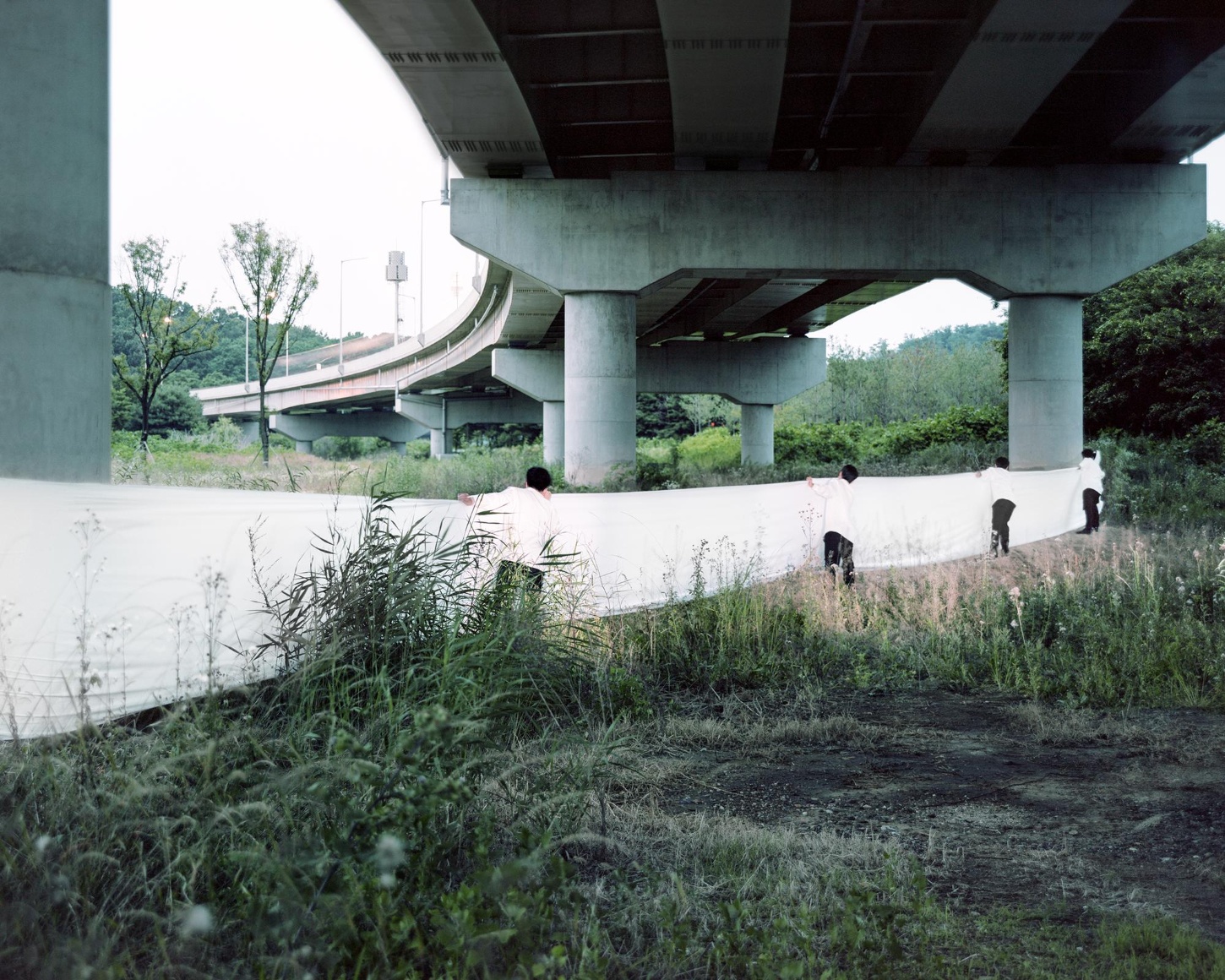 "Wandering, Wondering' by Seok-Woo Song — Beneath a multi-tiered elevated highway system, five uniformed figures occupy a concrete platform scattered with native vegetation and weeds.