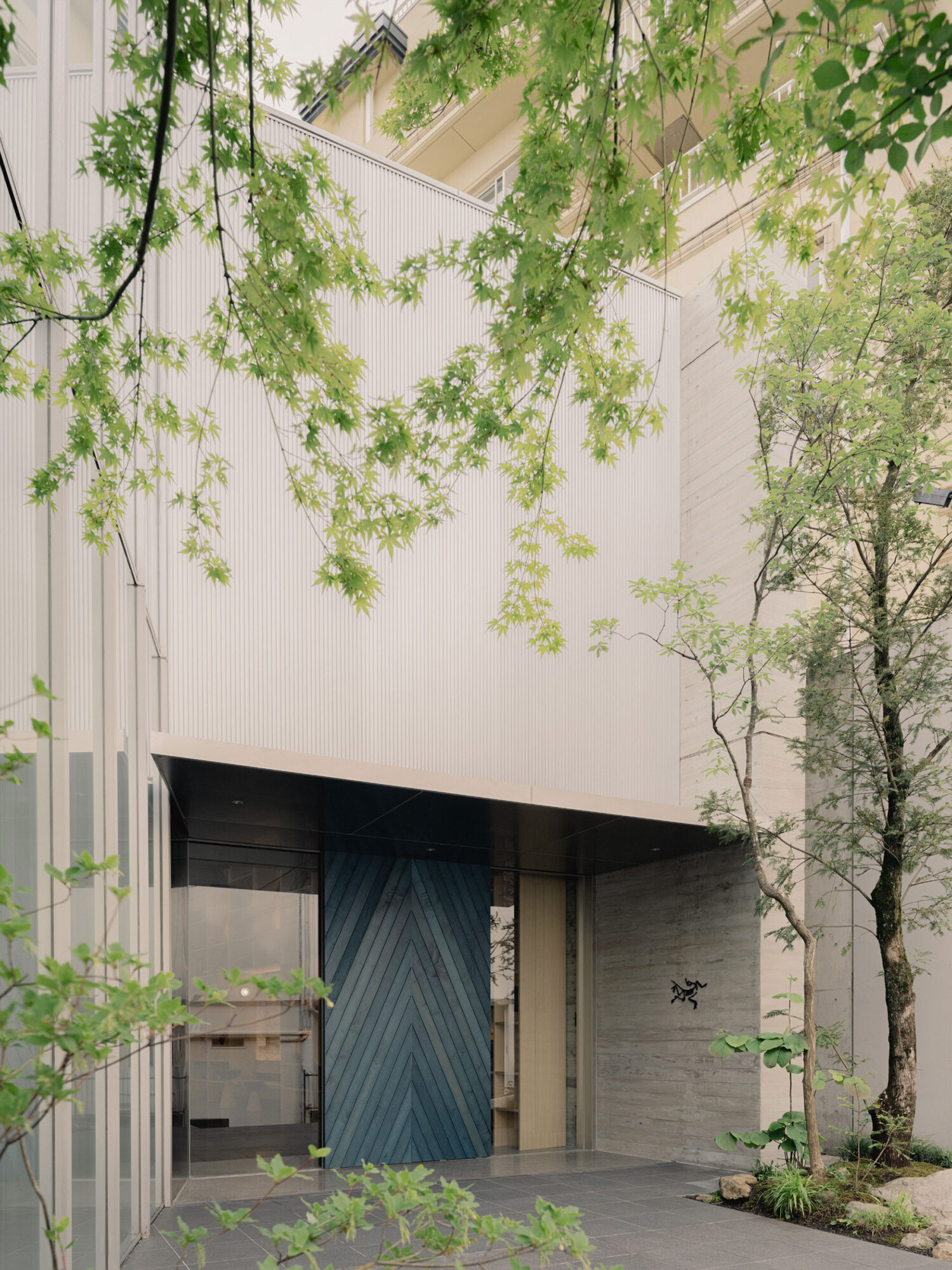 Arc’teryx Tokyo Creation Center by Torafu Architects — The entrance courtyard frames a geometric blue-grey timber door beneath a massive cantilevered concrete soffit with ribbed underside.