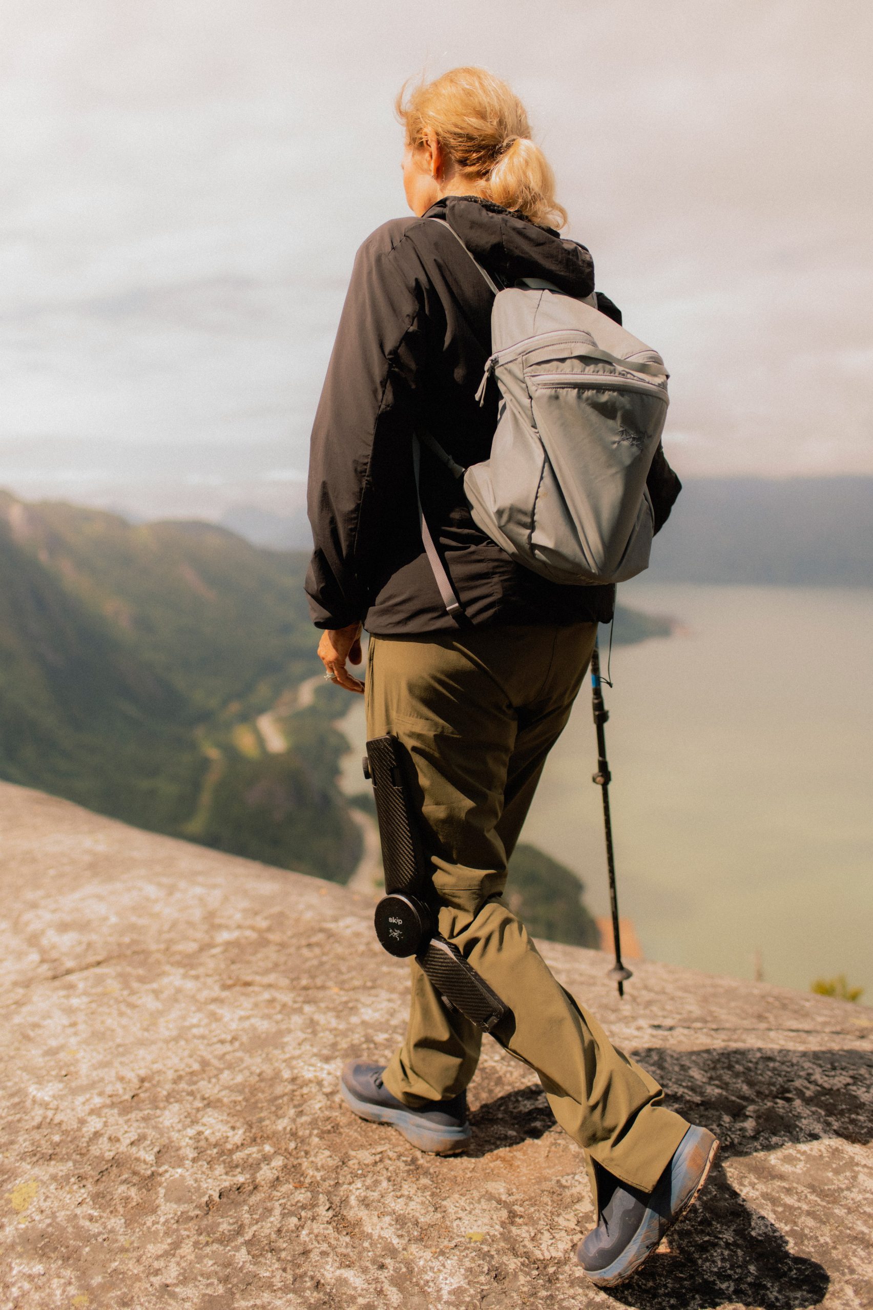 MO/GO Pants with Exoskeleton by Arc'teryx + Skip — A rear-facing photograph captures the wearer mid-stride on a clifftop overlooking a river valley, with the exoskeleton's mechanical components rendered...