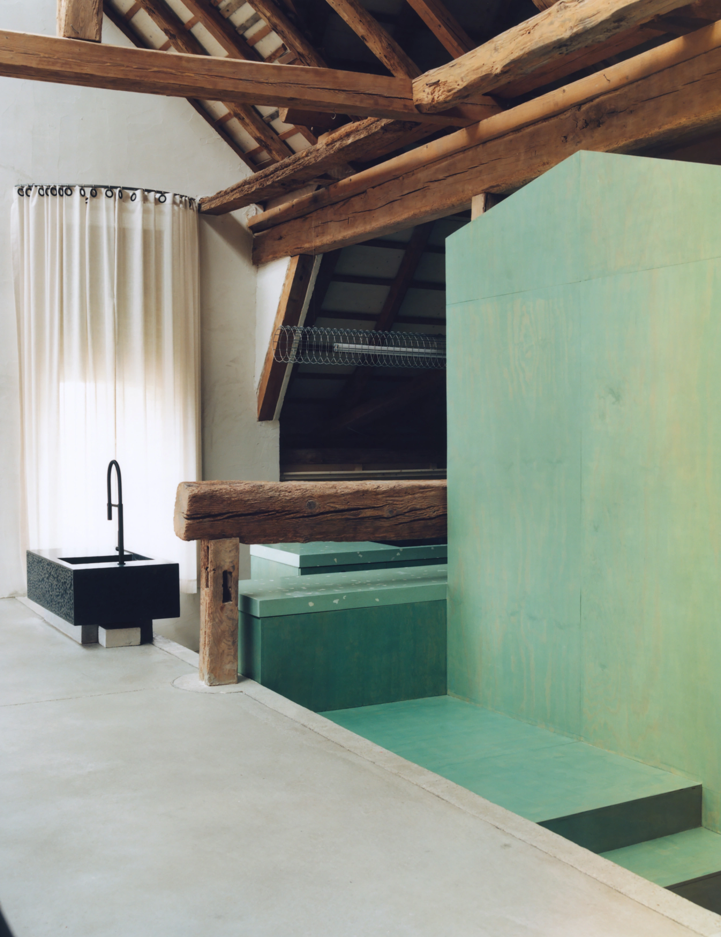 Erika and Willi's Barn by Piertzovanis Toews — A monolithic green platform anchors the right side of this barn interior, its stepped geometry creating integrated seating and storage.
