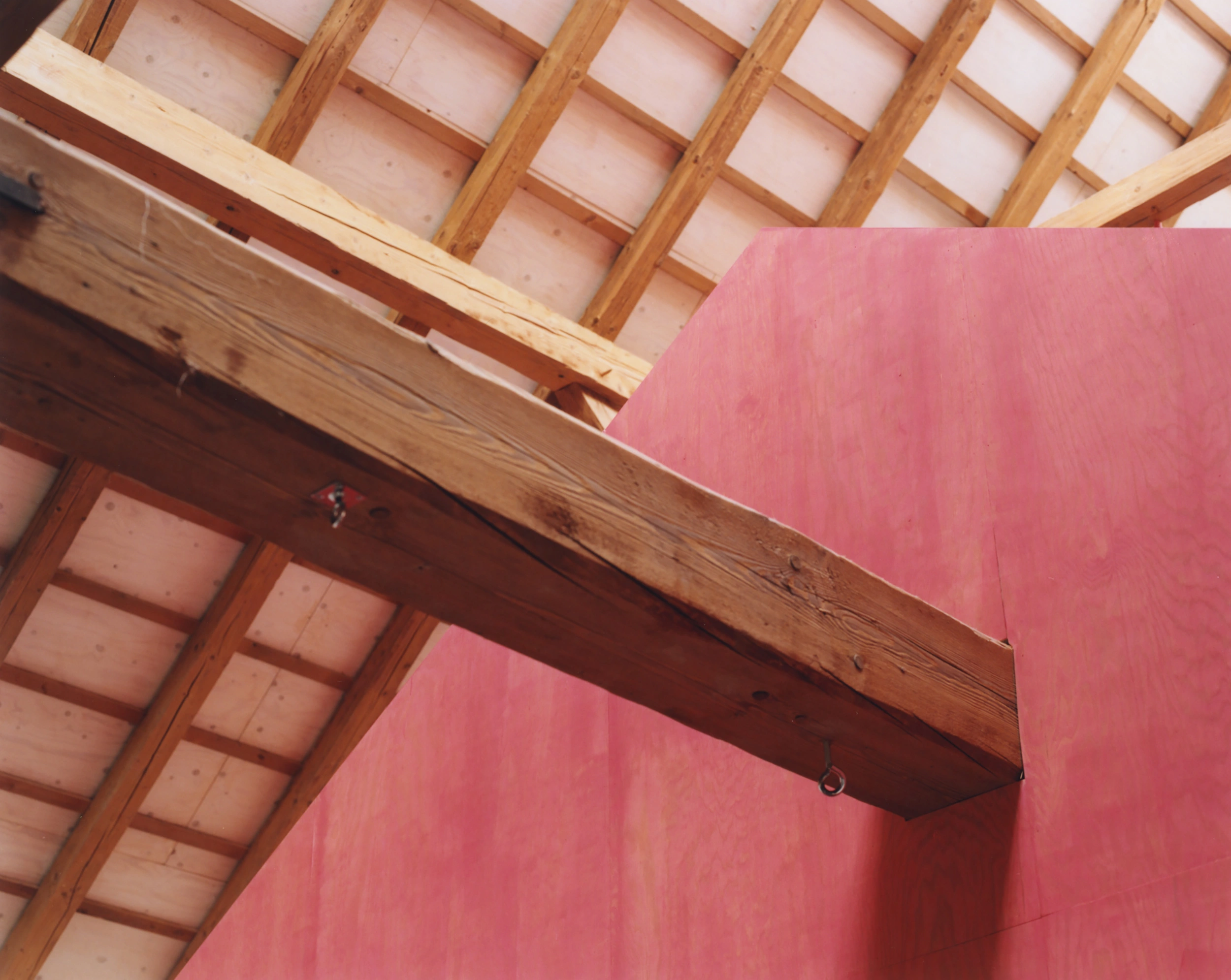 Erika and Willi's Barn by Piertzovanis Toews — A timber ceiling structure transitions from exposed coffered beams in warm honey tones above to a bold red-painted wall plane below.