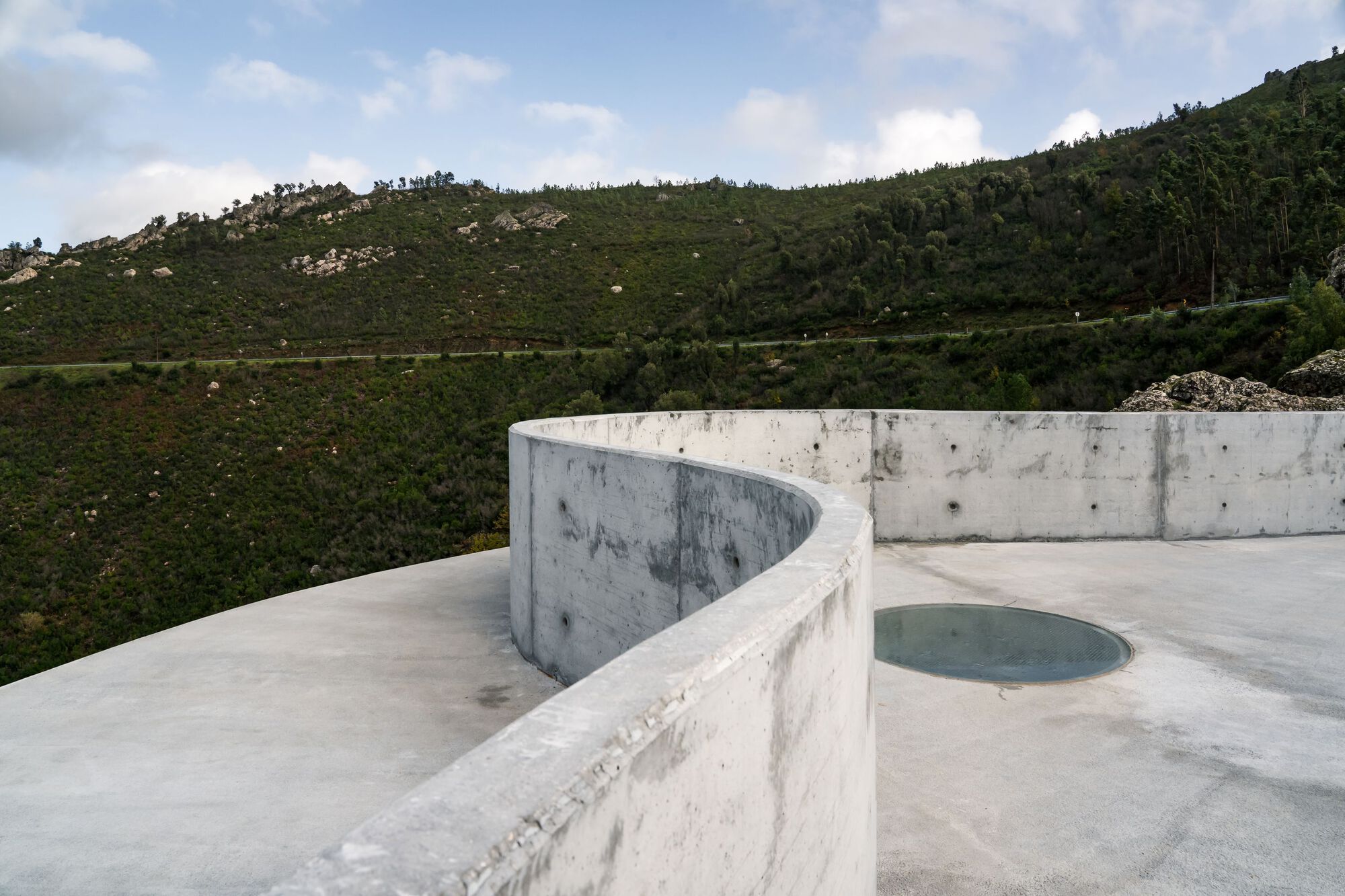 Renovation of the Zebro Viewpoint by Álvaro Siza Vieira by Alvaro Siza — A circular reinforced concrete platform cantilevers from a rocky stone base, its white monolithic form punctuated by an integrated circular glass panel...