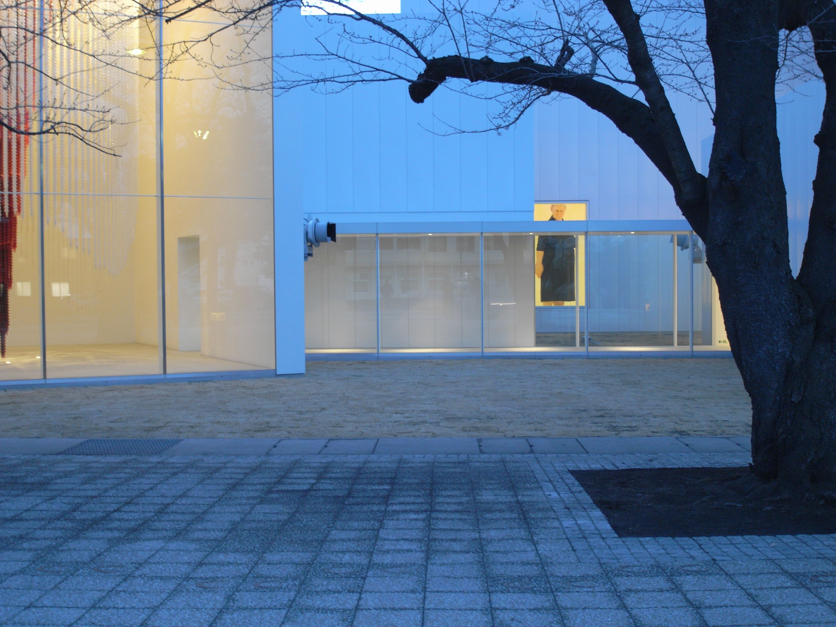Towada Art Center by SANAA — Twilight view of a pavilion's glazed facade framed by a bare tree branch.