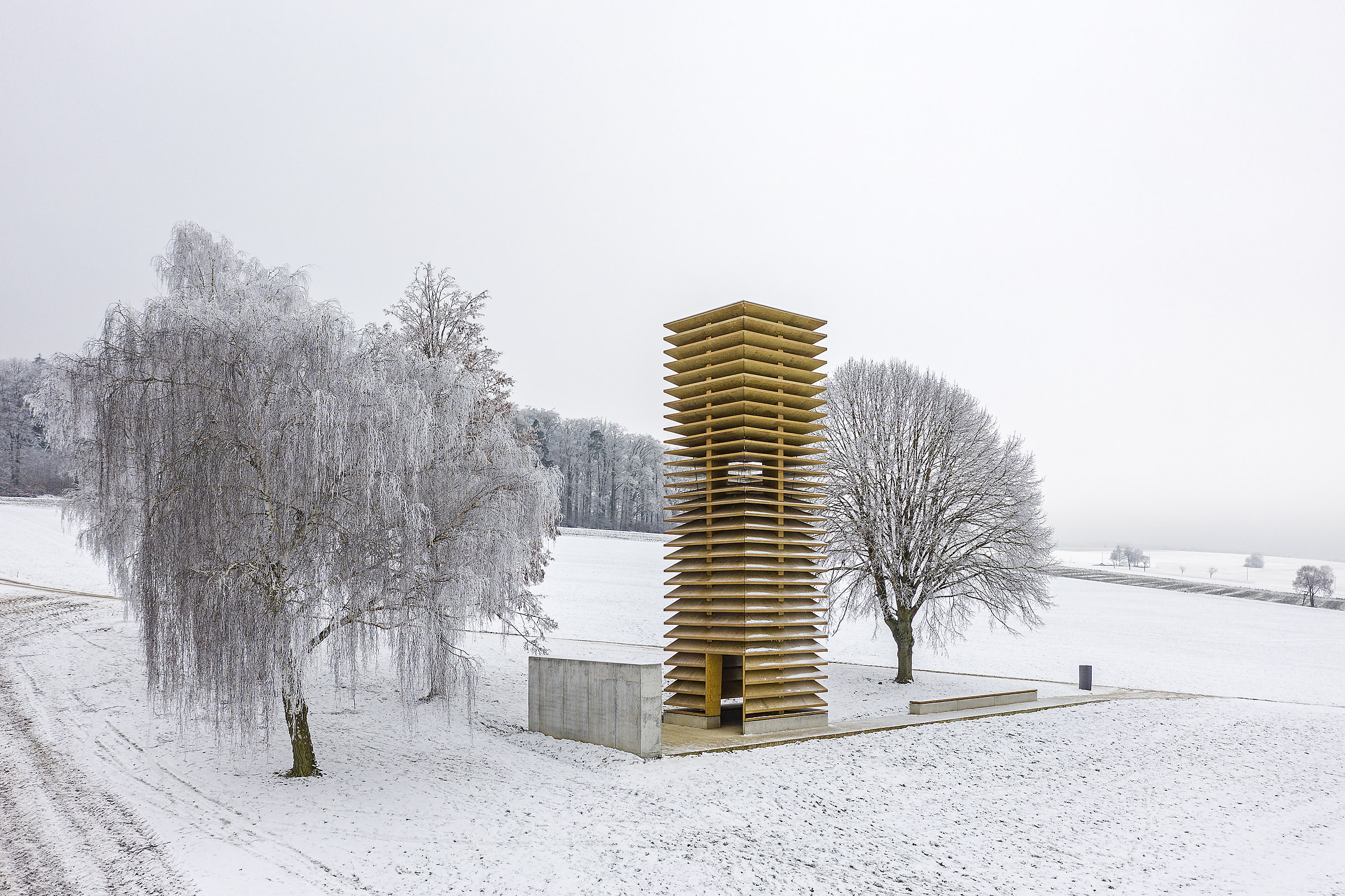 Kapelle (Chapel) Kesselostheim by Staab Architekten — A golden-brown timber tower rises from a geometric concrete platform set within a frost-covered field.